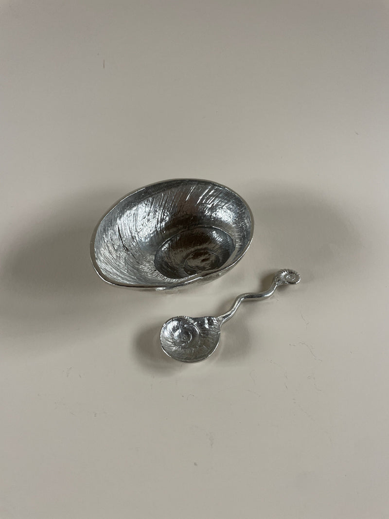 The Shell Dish & Spoon by Maud Vaughan, featuring a small, textured vintage polished pewter dish with a wavy design, sits beside its matching spoon on a plain light background.