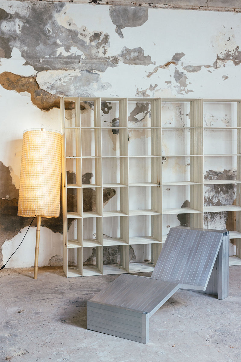 An Aluminum Lounge Chair by Studio HAOS sits beside empty wooden shelves and a tall floor lamp with a woven shade, all set against a weathered wall in an industrial-style room with modern accents.