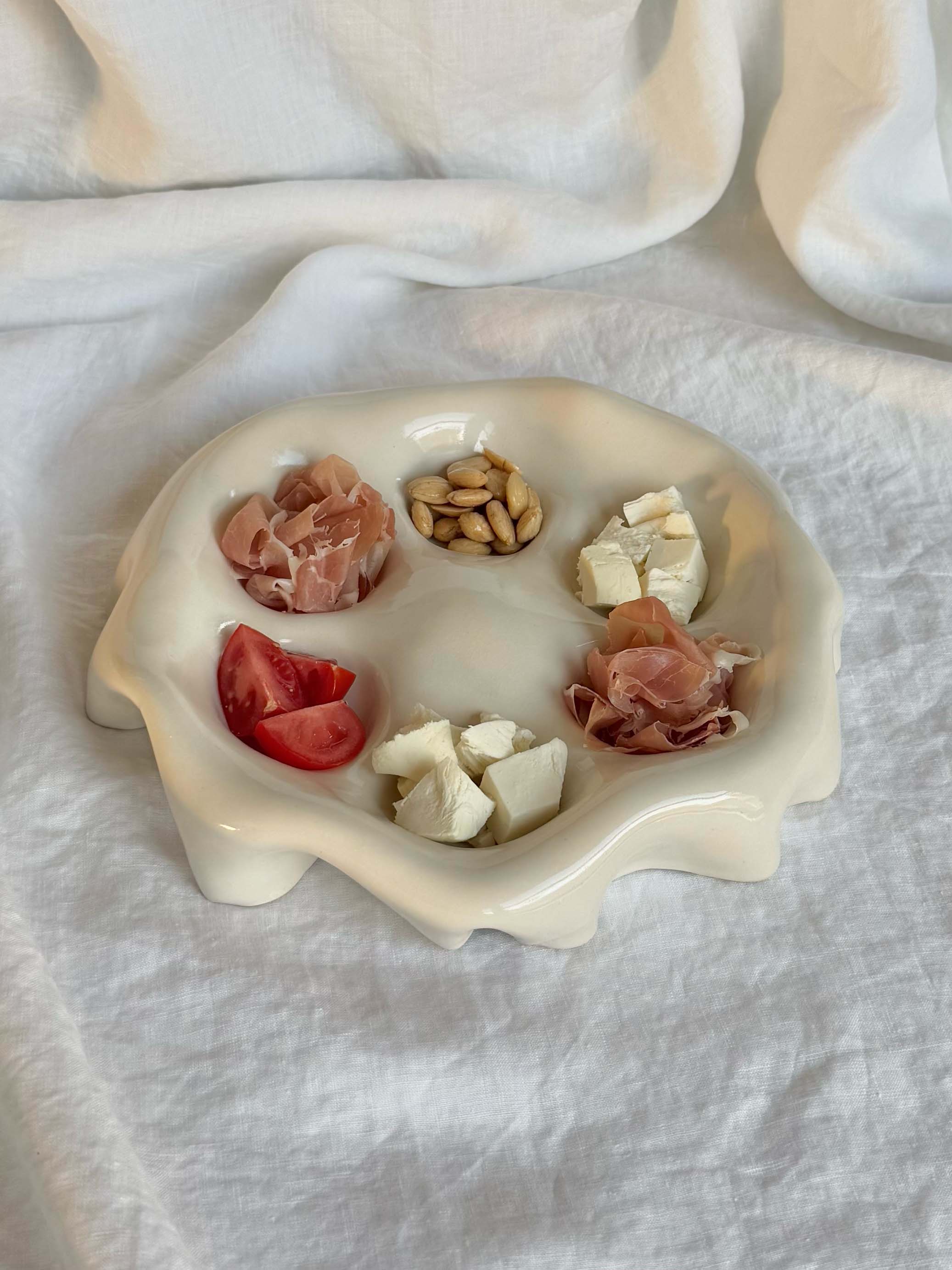 The Ceramic Snack Plate by Goobs Ceramics is a white dish made of hand-crafted stoneware clay, featuring five compartments for sliced prosciutto, pine nuts, tomato chunks, mozzarella pieces, and more prosciutto. Its high gloss glaze shines elegantly against a textured white fabric background.