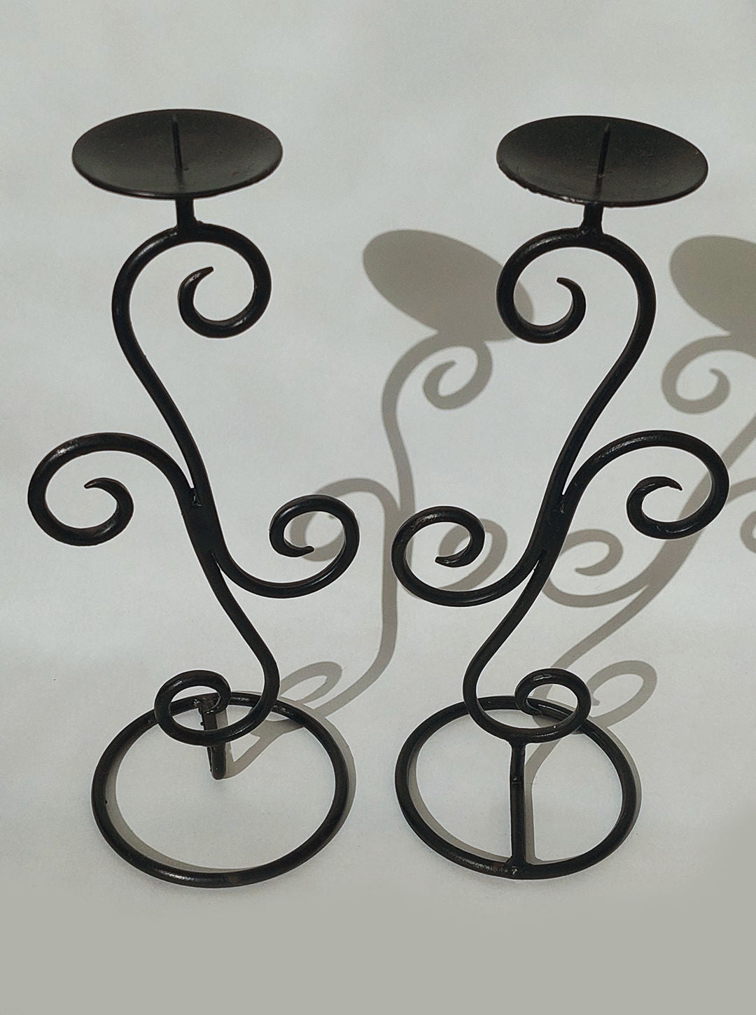 The Pair of Candle Holders by Les Objoies, crafted from black wrought iron with intricate spiral designs and rounded bases, stand side by side on a neutral background. Each holder features a flat disc atop for placing candles. Their curved shapes cast artistic shadows on the surface behind them, making them perfect home décor pieces.