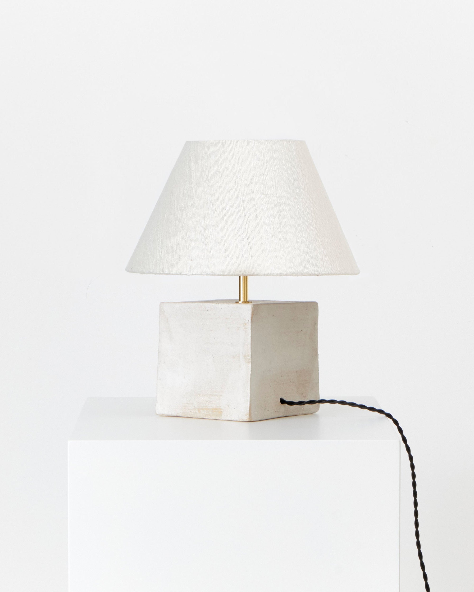 The Arouca Table Light White by Project 213A features a handmade square off-white ceramic base, paired with a white fabric lampshade and black cord—an artisanal, minimalist lamp showcased on a white pedestal against a plain background.