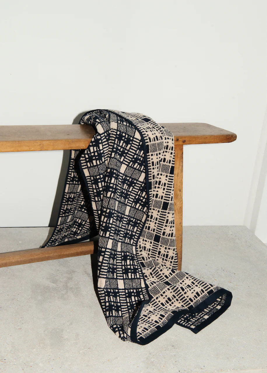 A black and beige checkered Alma Bath Towel by Autumn Sonata draped elegantly over a wooden bench against a plain white wall.