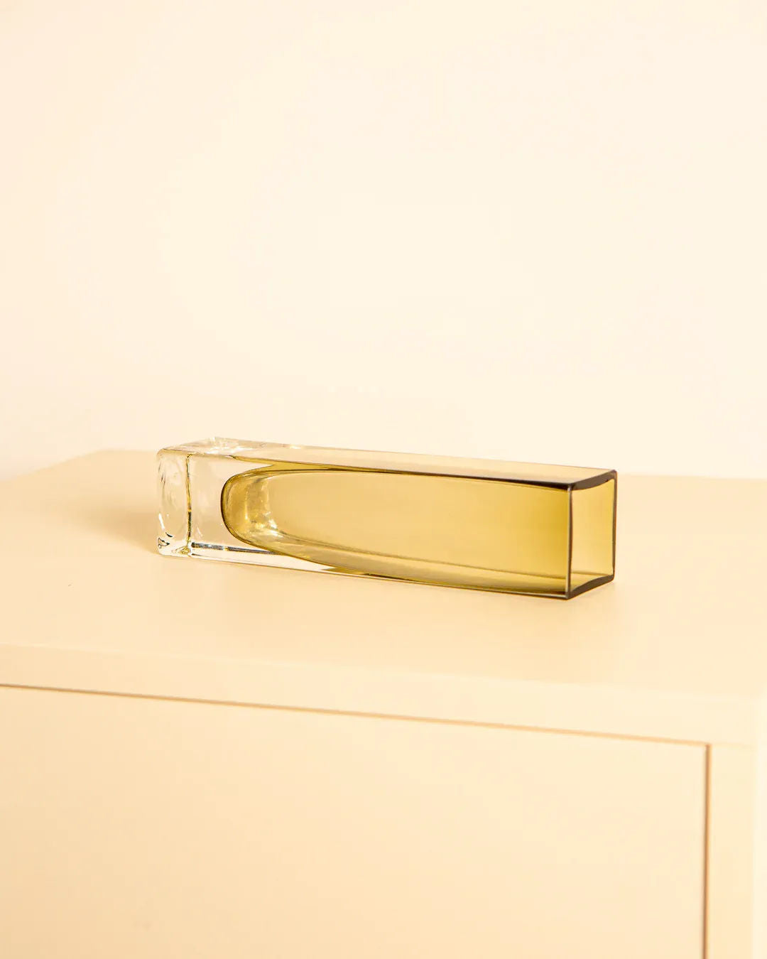 The 80's Murano Glass Amber Vase by Treaptyque, a tall, rectangular transparent glass bottle with a light brown hue, sits on a beige surface against a matching background. Its elongated design echoes the vintage elegance of Italian master glassmakers.