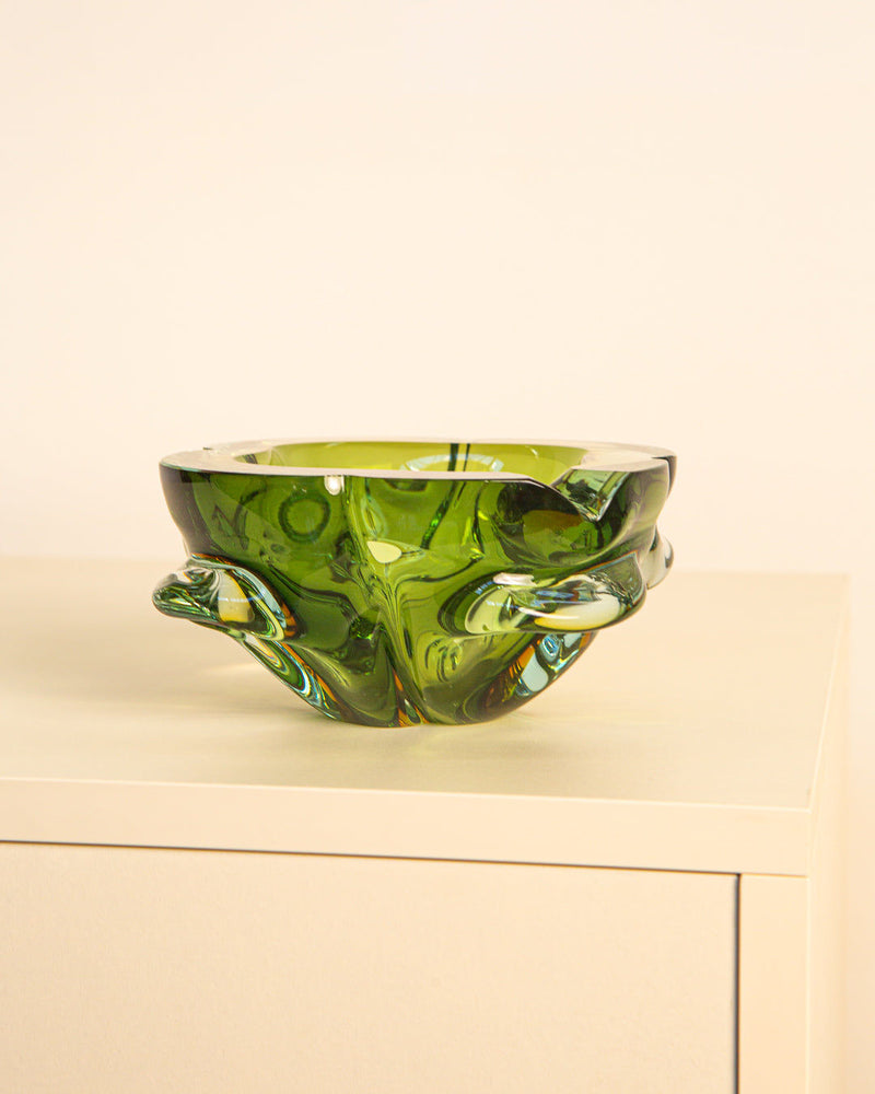 The Treaptyque Dark Green Murano Glass “Fleur” Ashtray from the 60s, with a wavy abstract design, sits on a light cabinet against a beige backdrop, embodying iconic Italian style.