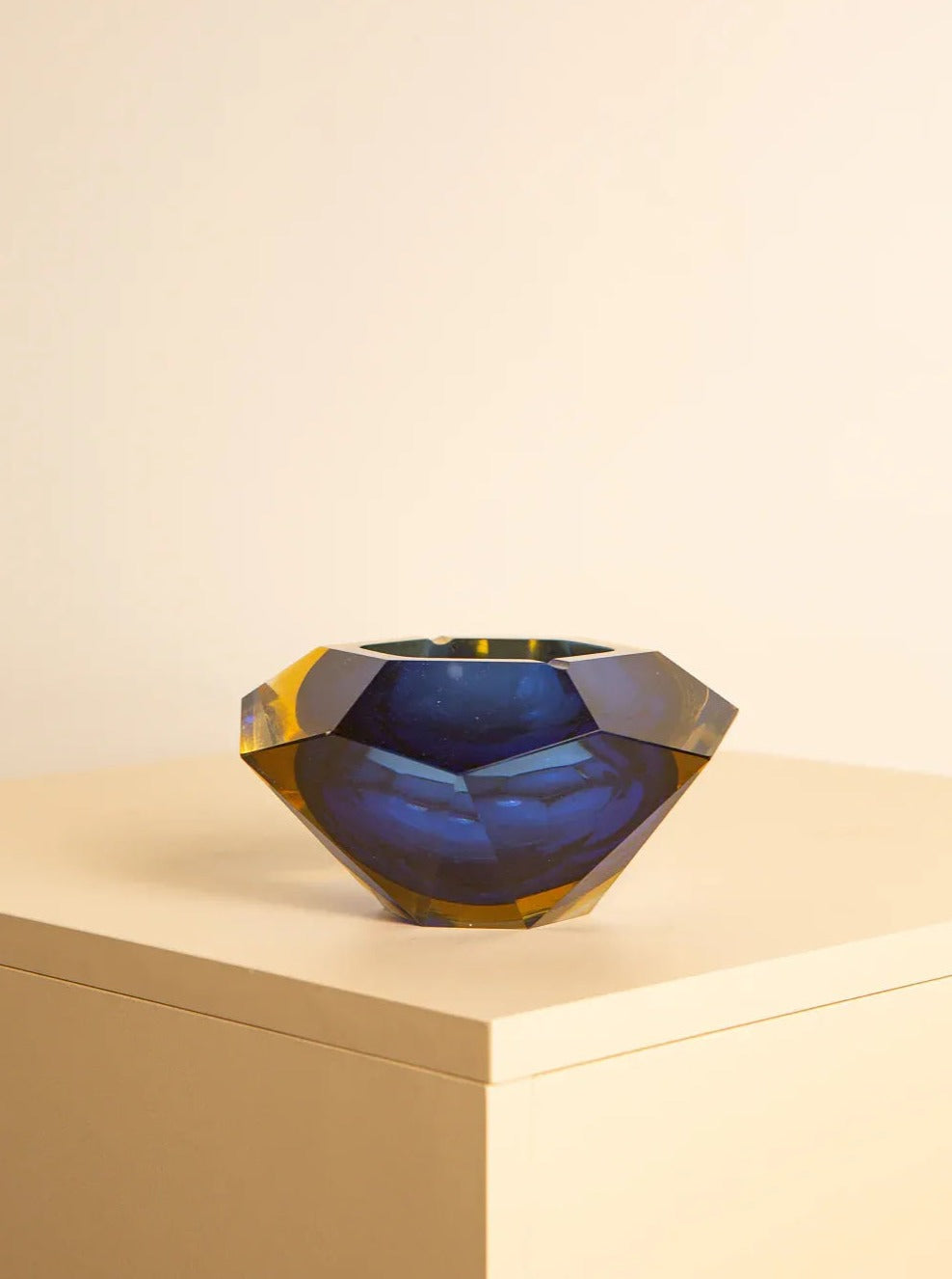 A Large vide-poches blue "Diamond" by Flavio Poli 60's from Treaptyque with a deep blue center and yellow edges, displayed on a light-colored surface against a beige background. This handcrafted piece of Italian glass art features an angular design and rich colors that create a striking visual contrast.