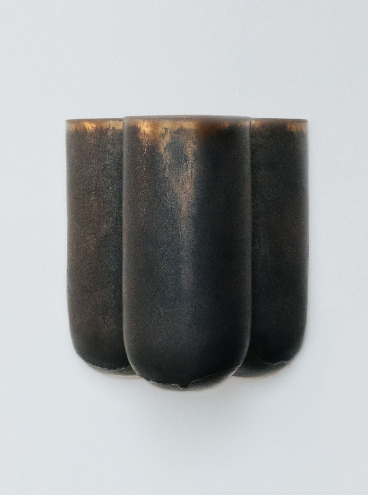 Introducing the Wall lamp MOOR PLUS BRILLANCE by Lisa Allegra: a minimalist, dark metallic wall sconce featuring a three-lobed, sculptural design. The light fixture has a matte enamel finish, offering a sleek and modern appearance against the plain white background.