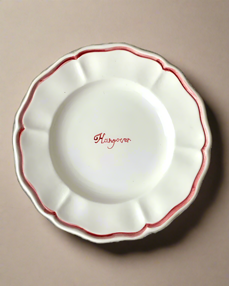 The "Hangover" Fil Rouge Plate by Musae Studio is a decorative white plate with wavy edges and a red trim, featuring the word "Hangover" in red cursive font at its center. Set against a textured green surface, this plate serves as the perfect party starter.