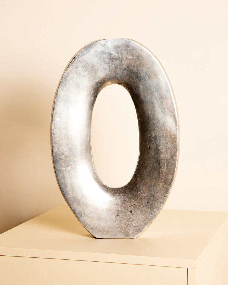 The Treaptyque Large Cast Aluminium “Oval” Vase, inspired by 80s Italian craftsmanship, features a metallic finish and hollow center, elegantly displayed on a beige surface against a neutral background.