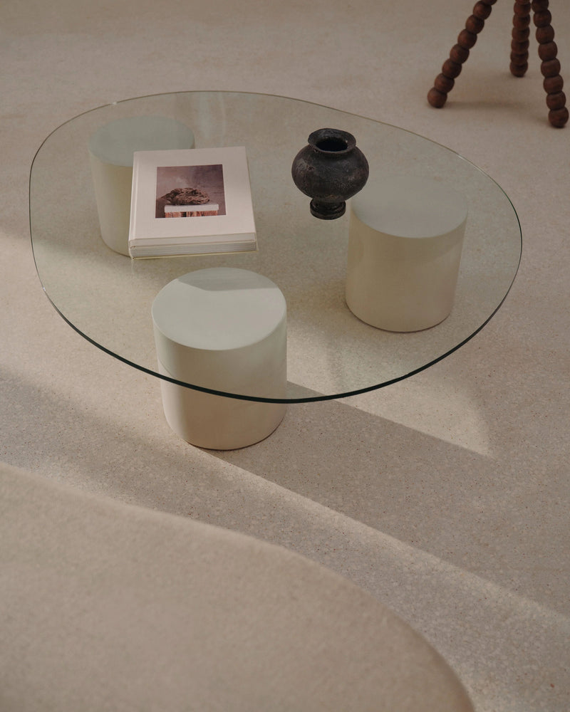 A minimalist living room scene showcases the "Mattia" Glass Coffee Table by MarlotBaus, featuring two cylindrical supports. The table holds a stack of books and a small, dark vase. Microcement floors complement the soft, neutral-toned decor as sunlight casts gentle shadows across the space.