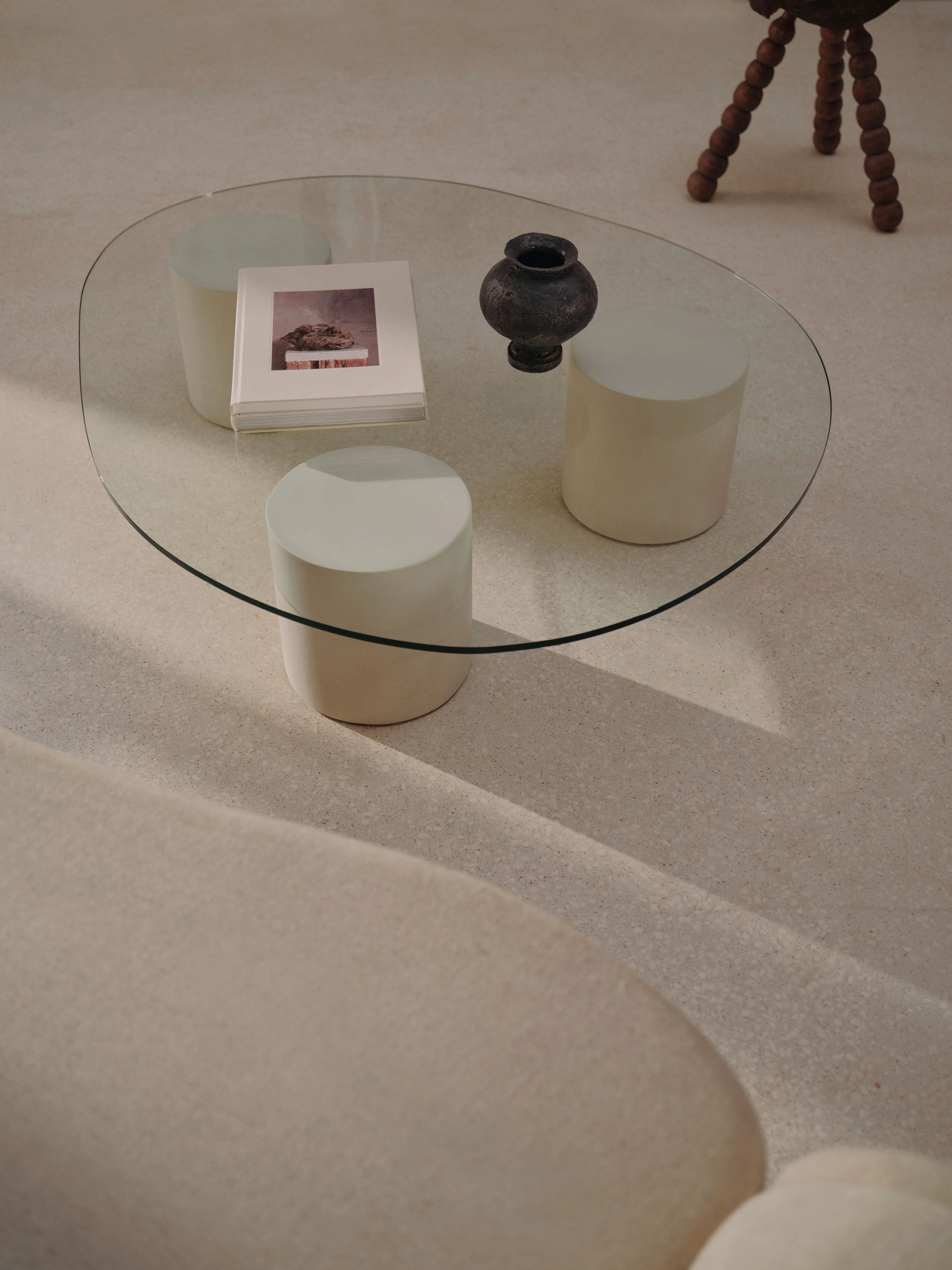 A minimalist living room scene showcases the "Mattia" Glass Coffee Table by MarlotBaus, featuring two cylindrical supports. The table holds a stack of books and a small, dark vase. Microcement floors complement the soft, neutral-toned decor as sunlight casts gentle shadows across the space.