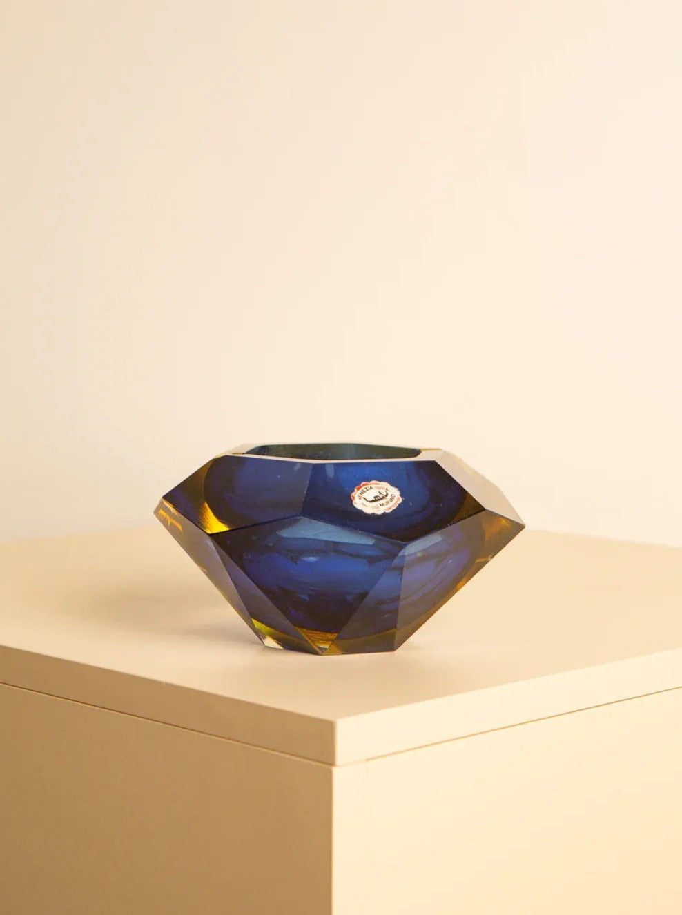 A Large vide-poches blue "Diamond" by Flavio Poli 60's from Treaptyque, faceted and deep blue with slight amber tones around the edges, shaped like a gemstone, sits on a cream-colored pedestal. Handcrafted in the tradition of Italian glass art, it features a small, round, rose gold emblem attached to its side. The background is beige.