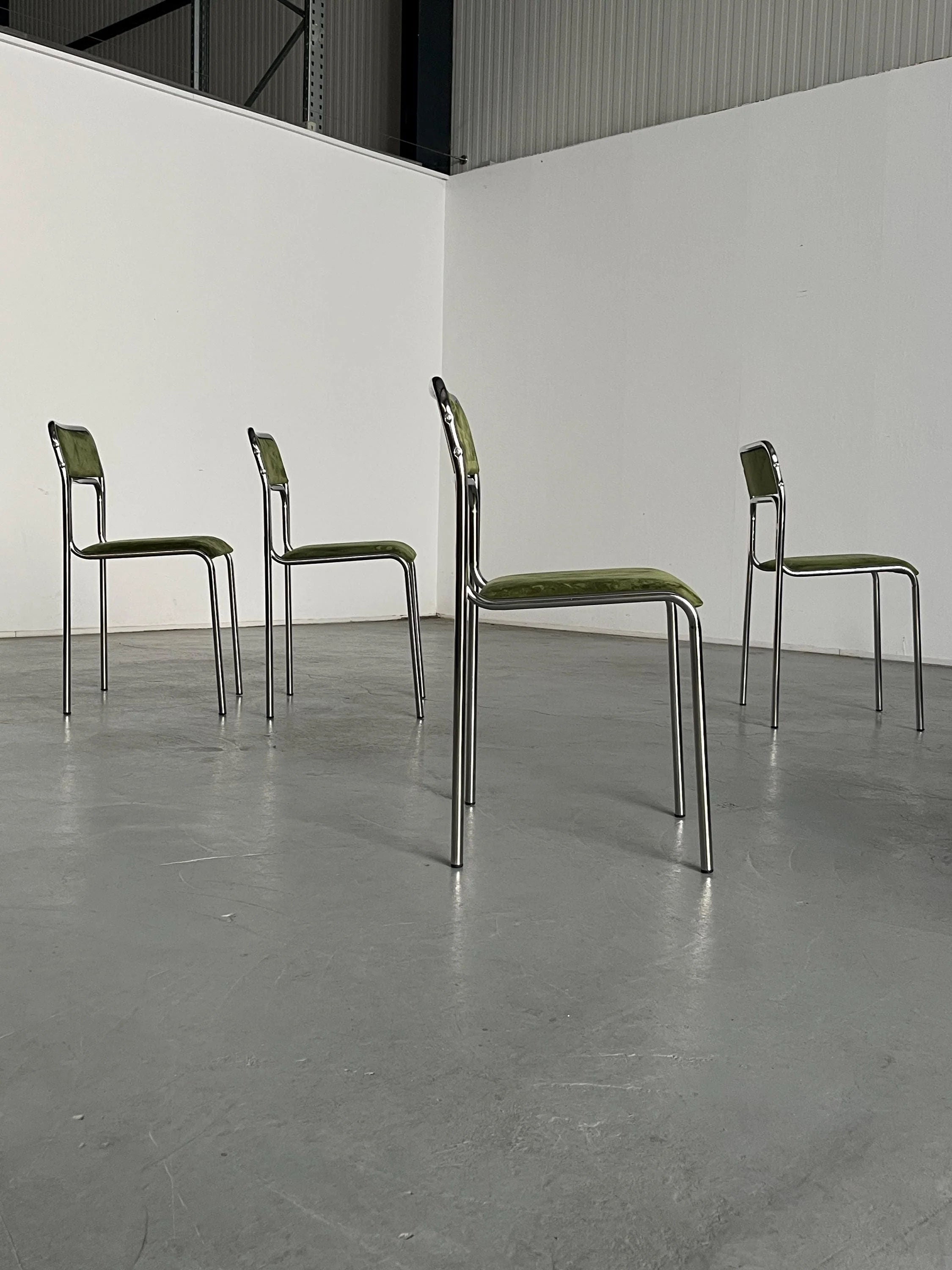 In a spacious industrial-style room with gray flooring and white walls, four Cherry Cargo Bauhaus chrome and green velvet chairs from a set of six are arranged in a square with one chair on each side facing inward.