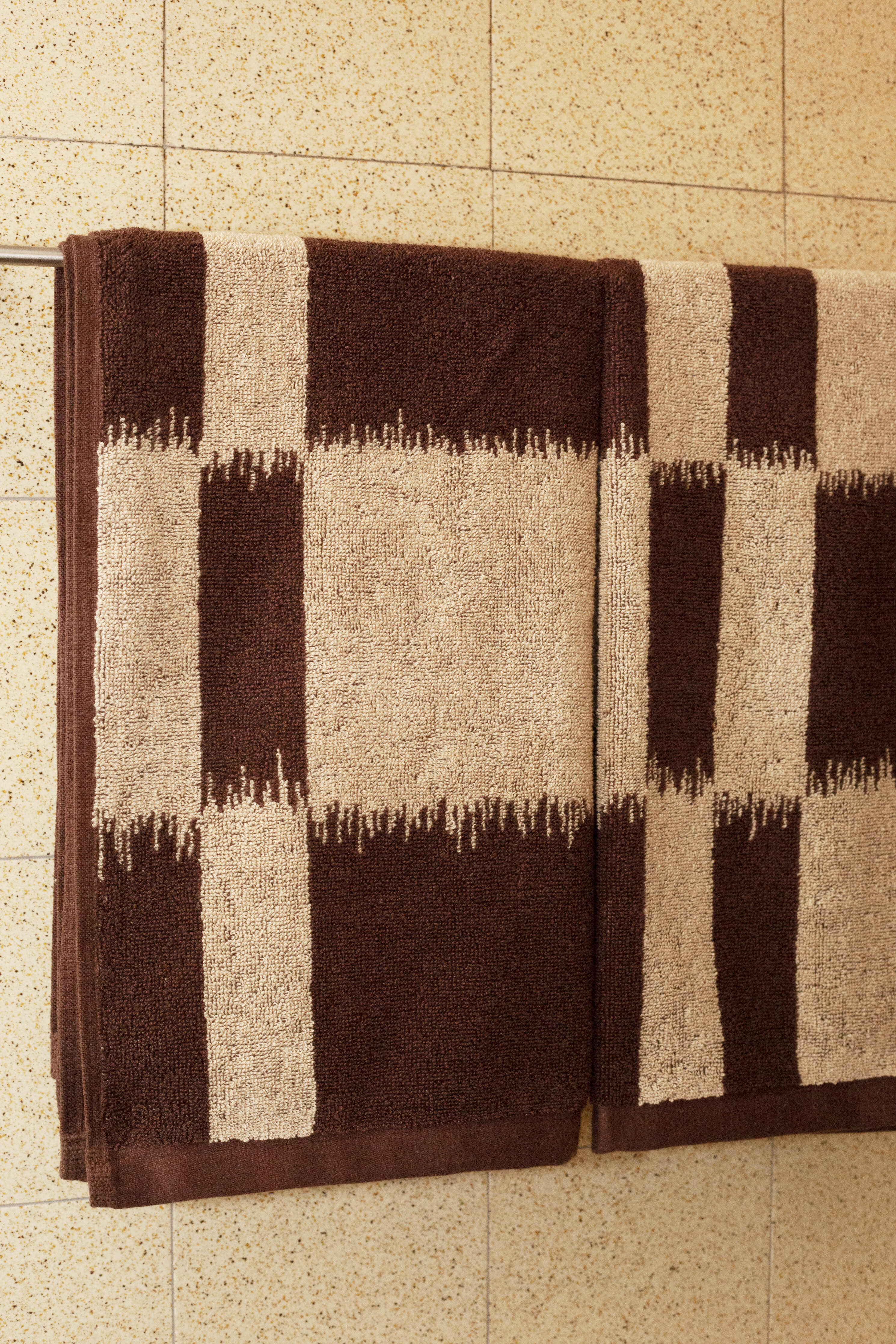 Two Karin hand towels with a brown and beige Ikat pattern hanging on a wall-mounted towel bar, set against a tiled beige background.