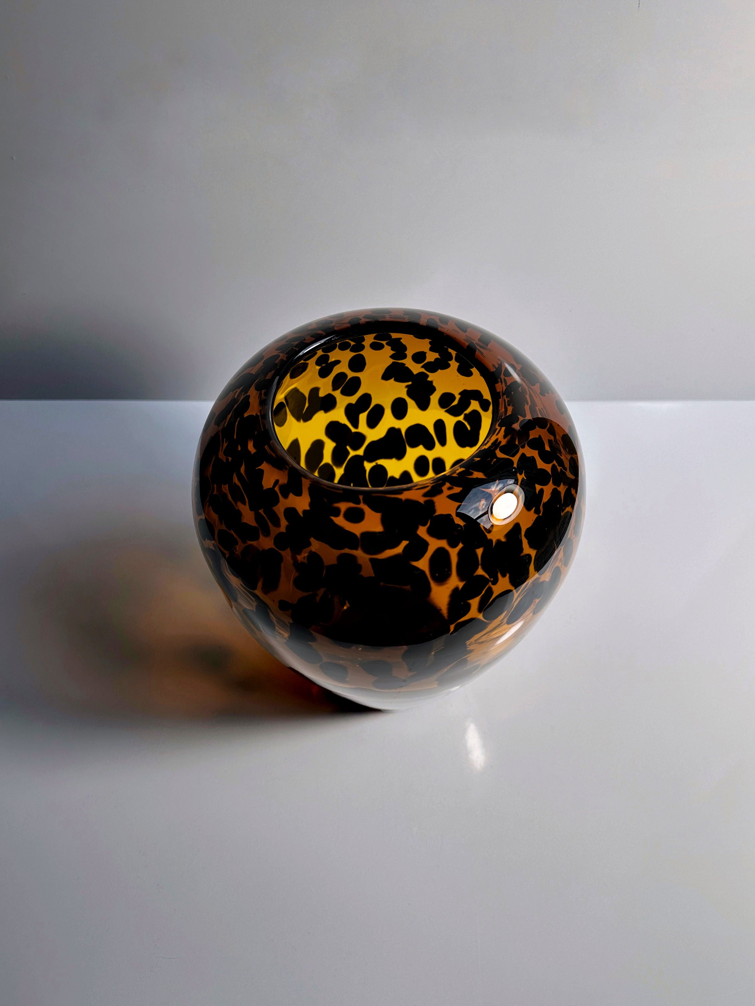 The Tortoise vase Murano for Christian Dior from the 1960s, offered by Dodo Vintage, showcases a round glossy glass design with a leopard print pattern of black and brown spots on a gold background. Placed on a light-colored surface, it exudes timeless elegance reminiscent of Christian Dior while casting a shadow to the left.