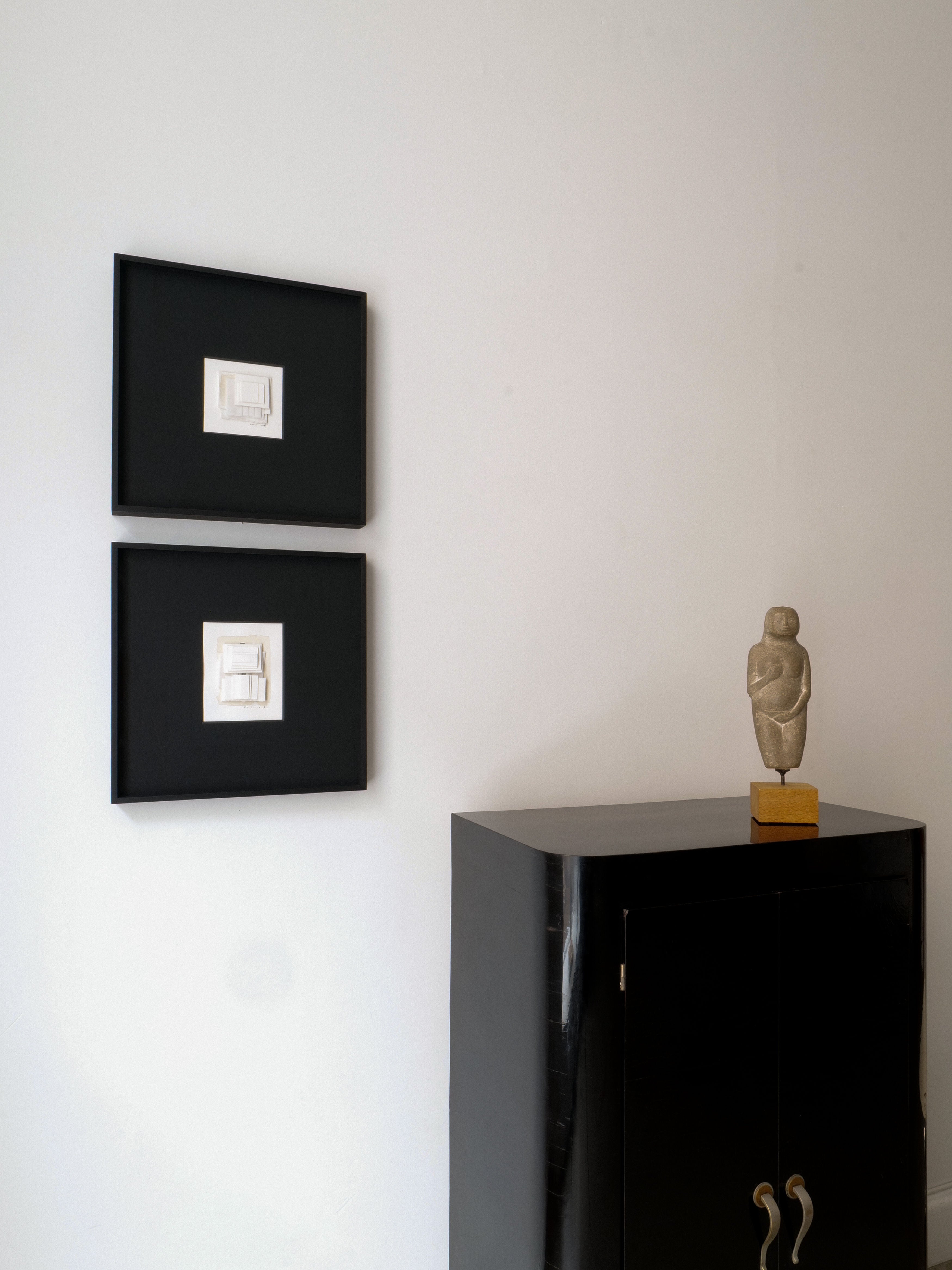 A minimalistic room features two framed artworks on a white wall above a black cabinet. A serene Marble Figurine Sculpture by Håkan Bonds from Collection apart stands on the cabinet, adding an artistic touch to the clean and modern decor.