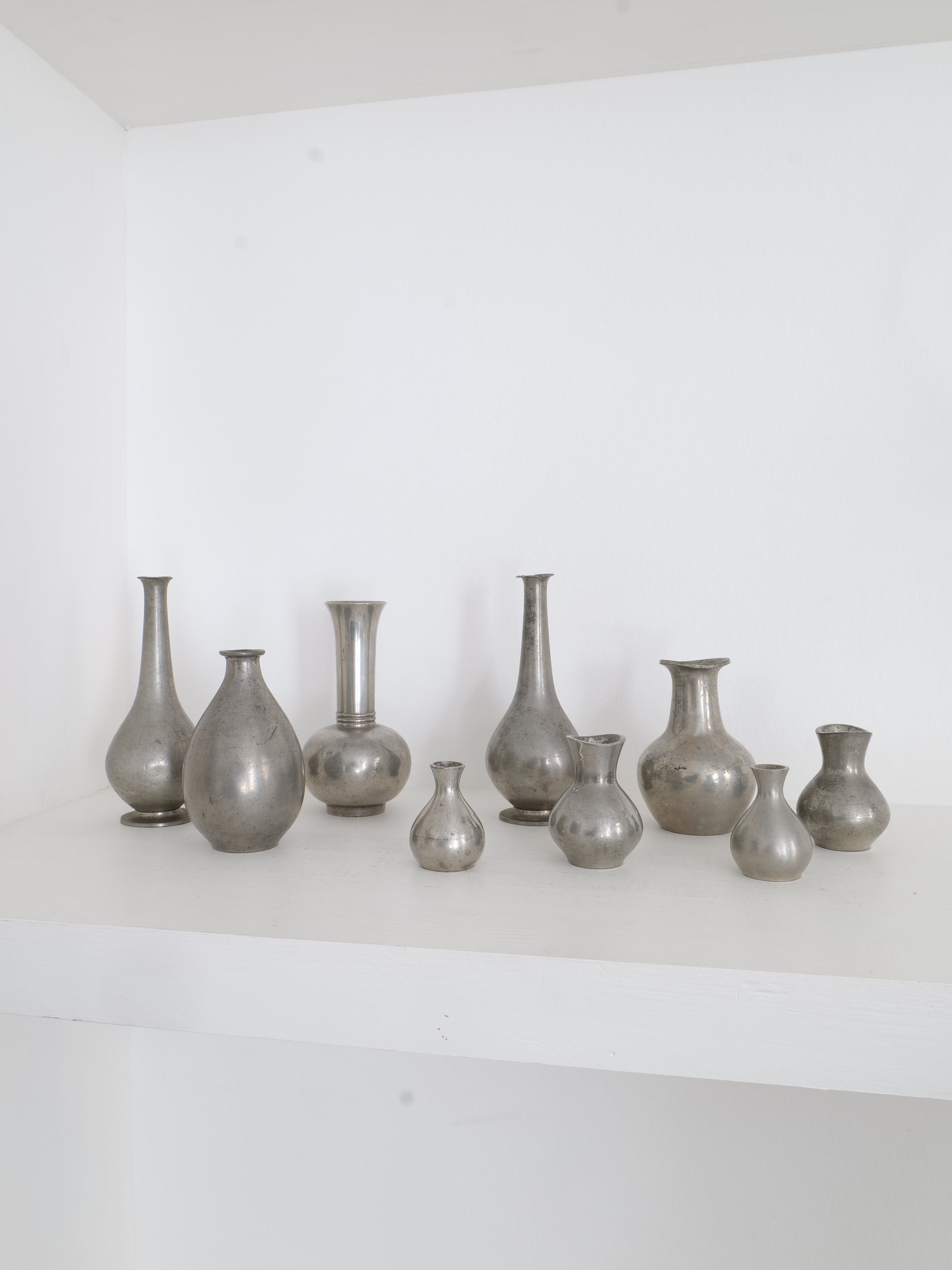 A collection of the Just Andersen Mini Vase Collection by Collection apart is displayed on a white shelf against a white wall. The small, metallic vases, reminiscent of the work of Danish silversmith Just Andersen, are arranged in various shapes and sizes. Their reflective surfaces and smooth contours are showcased in an aesthetically pleasing manner.