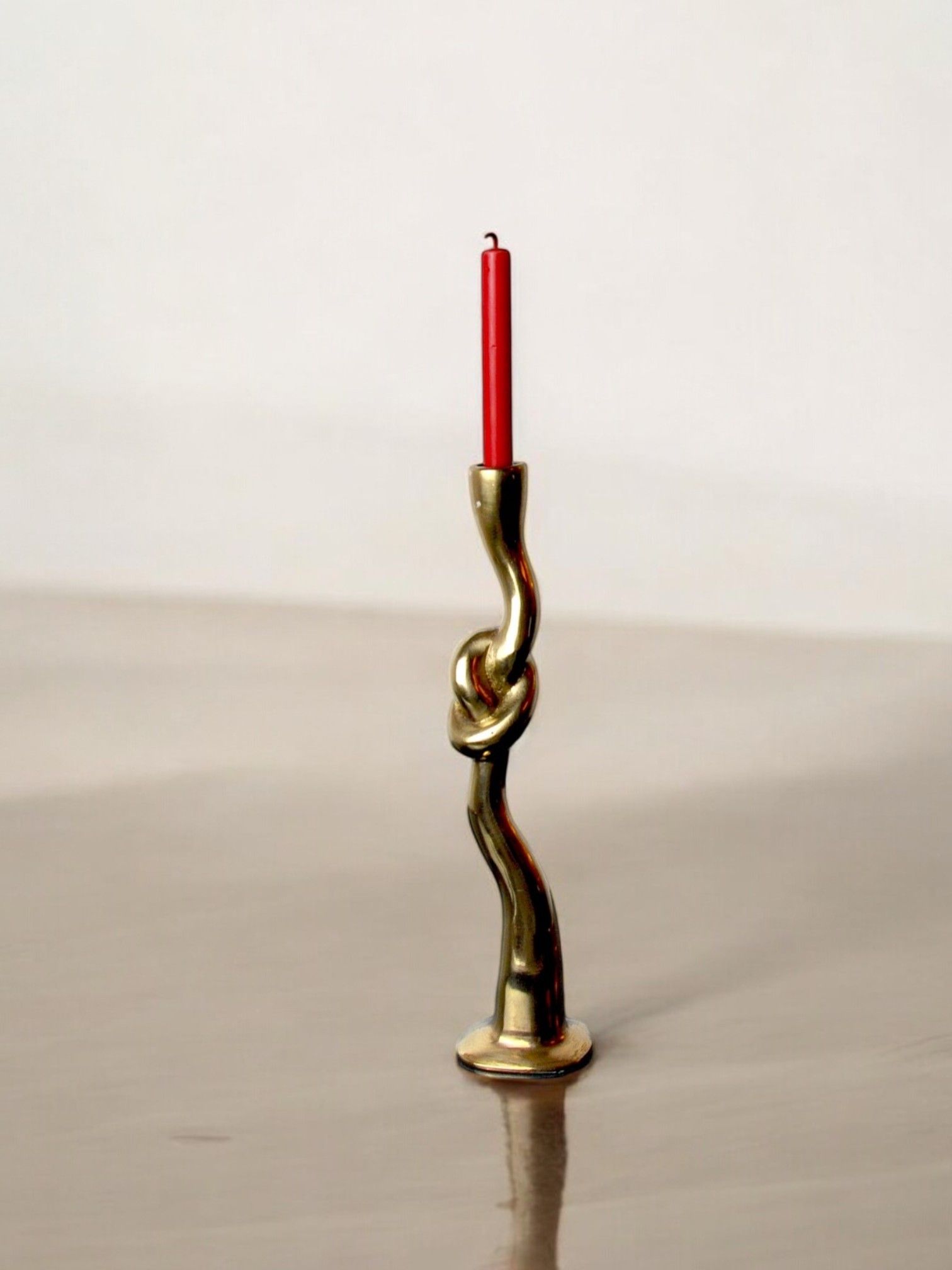 The Brutalist Knot Candleholder in Bronze 70s from Malamar Studio, featuring a twisted and smooth abstract design, holds a tall, unlit red candle. The background is a neutral, blurred surface.