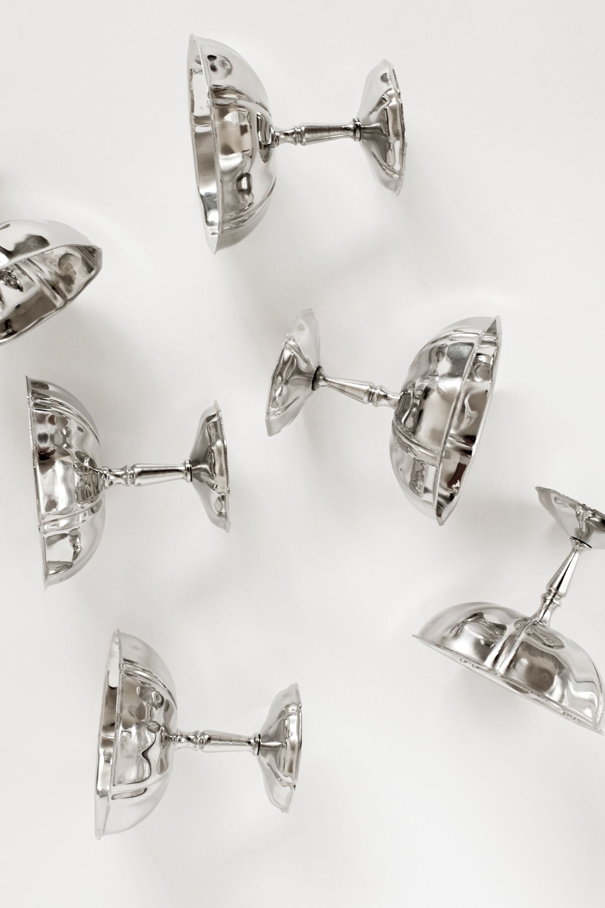 A Set of Six Ice Coupes by septembre studios is arranged upside down and sideways on a white surface, showcasing their hammered texture that reflects light and shadows, giving these elegant pieces a distinctive vintage charm.