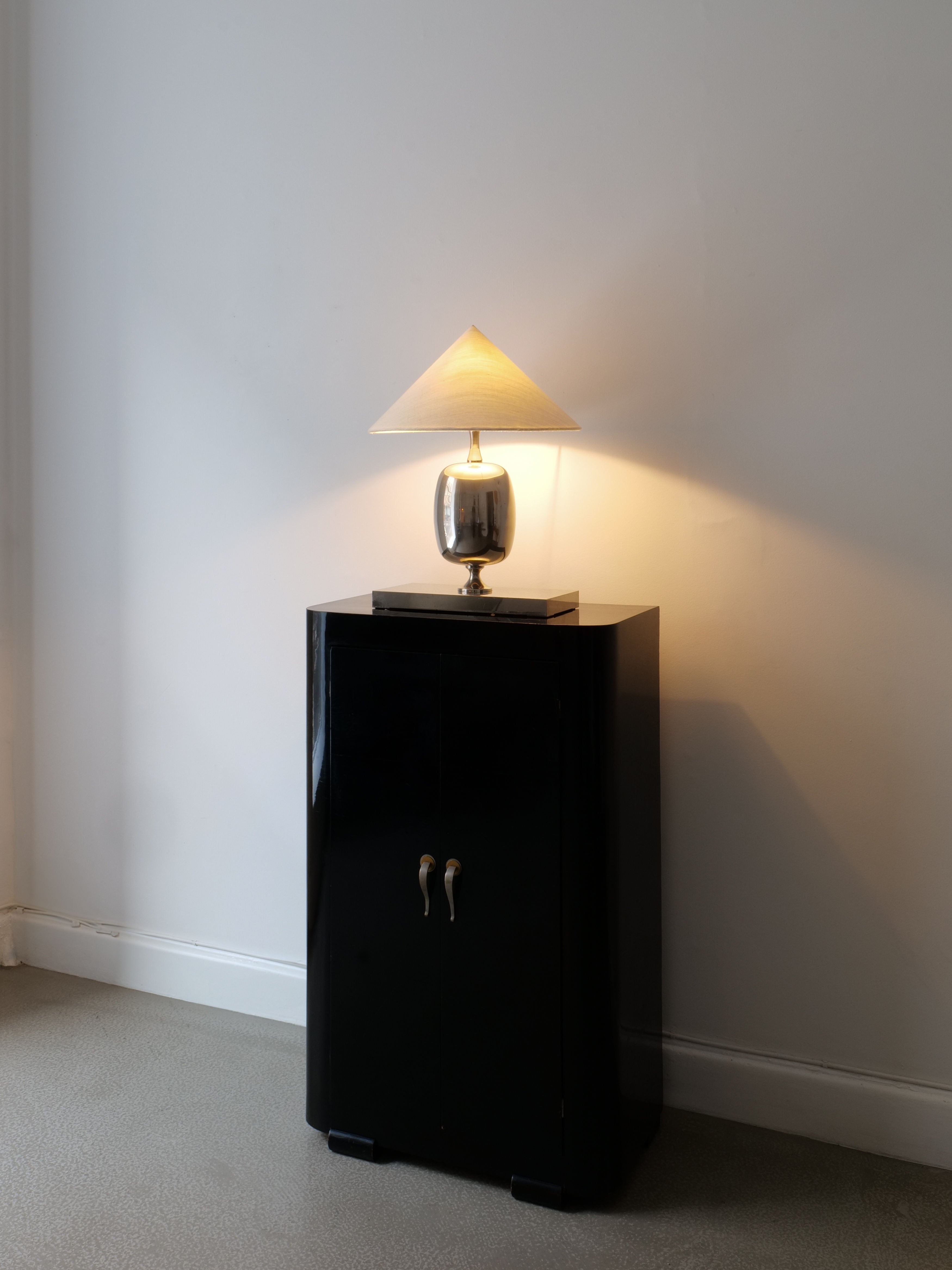 A small black cabinet with two doors stands against a plain white wall. On top of the cabinet is the Barbier Chrome Table Lamp by Collection apart, its conical lampshade casting a warm glow. The room has a light gray carpet.