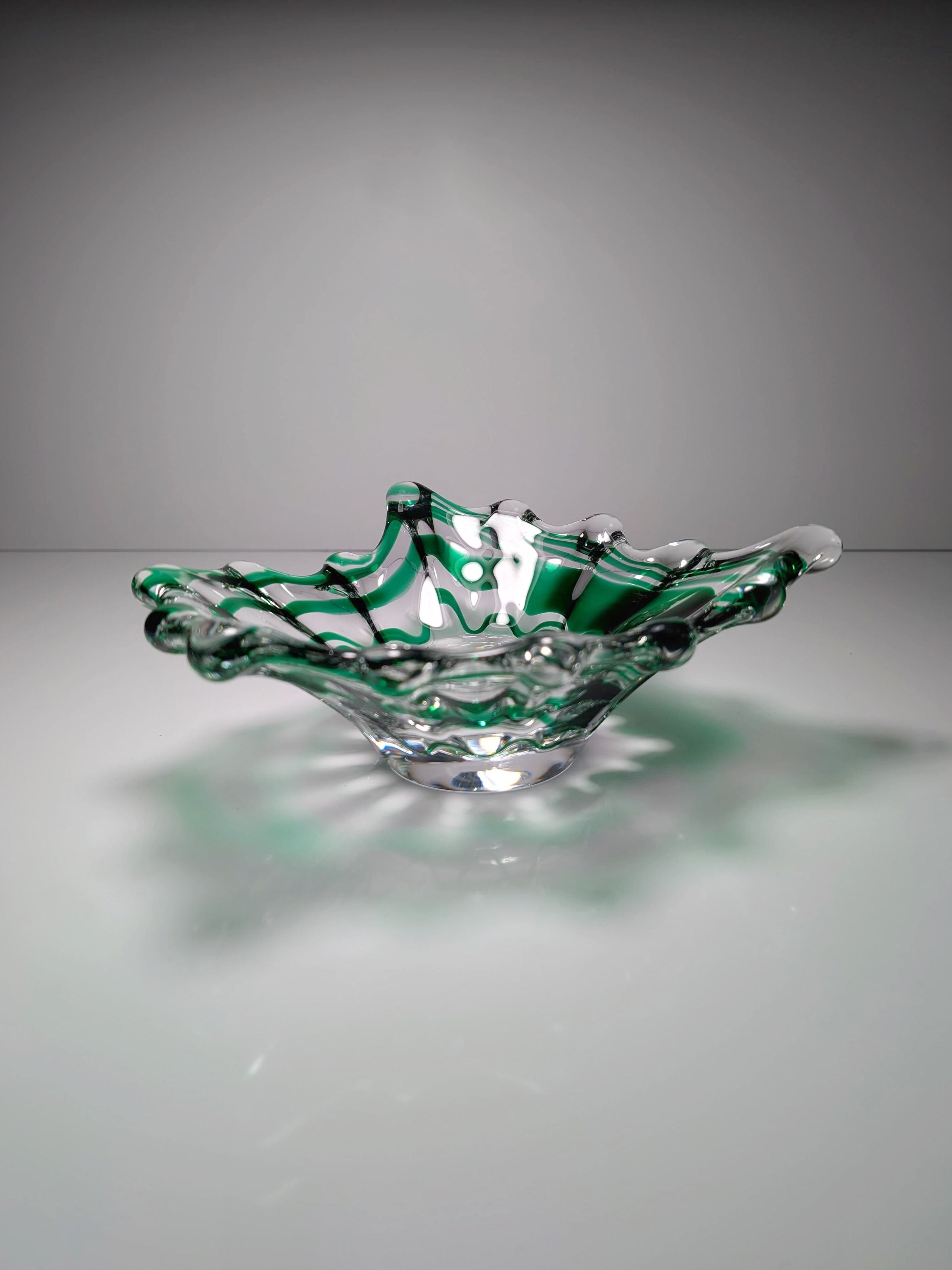 A Dodo Vintage Green Swirled Murano Vide-Poche sits on a reflective surface, its transparent wavy design and green and white swirls casting soft shadows that highlight its intricate details.