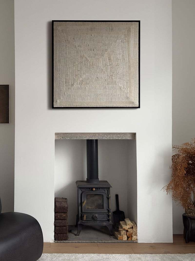 A minimalist living room showcases a black wood-burning stove nestled in a white alcove, enhanced by the rustic charm of solid oak accents. Above the stove hangs "Mending II, 2024," an artwork by Bec Kirby, while firewood and tools are neatly arranged nearby. To the right side of the alcove, dried plants introduce a natural element.