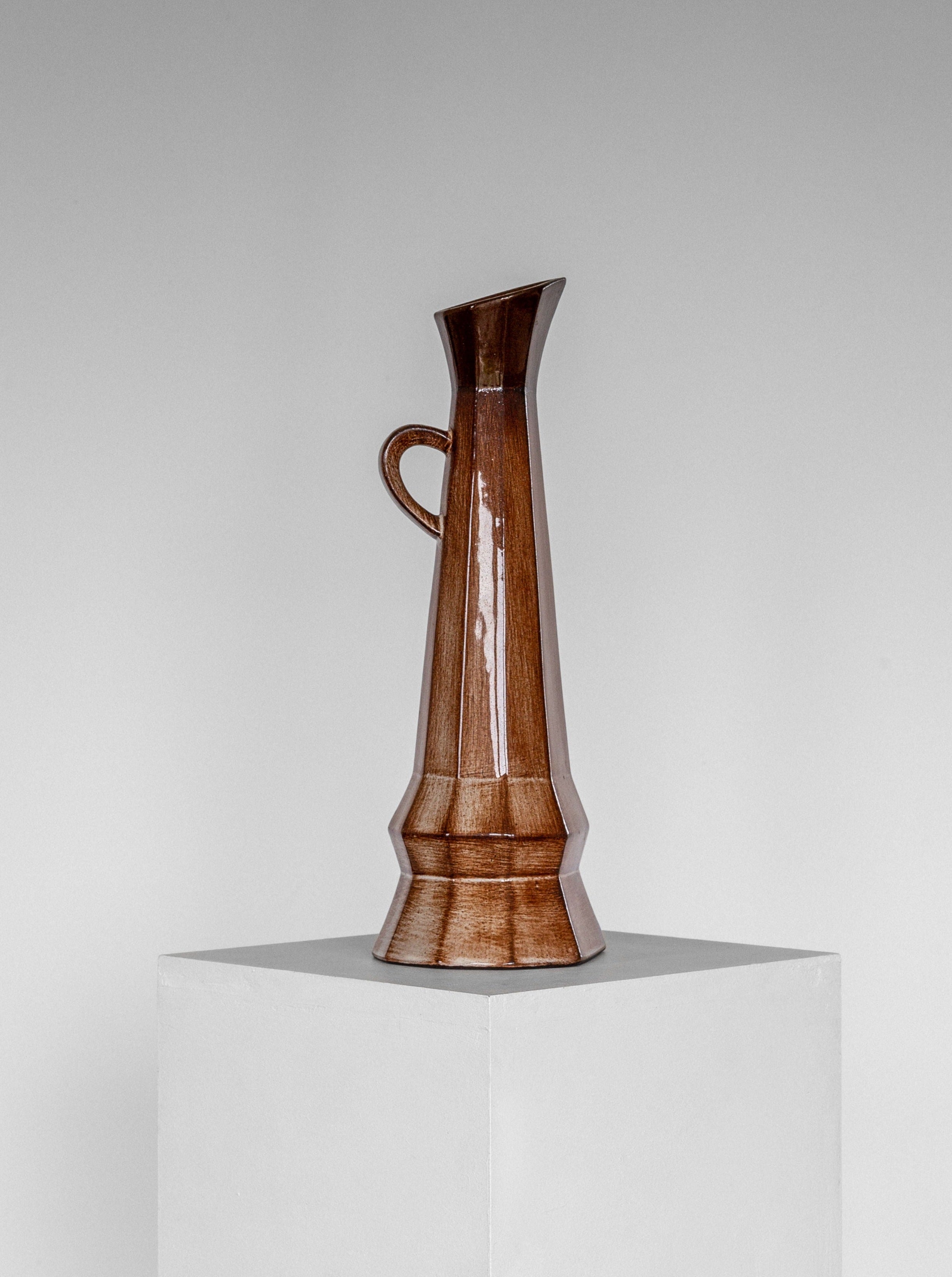 A tall, slender Cubist Ceramic Pitcher by Spigel, featuring a small handle near the top and a glossy brown finish with geometric facets, is displayed on a white pedestal against a plain gray background. This mid-20th-century vase has a slightly tapered spout.