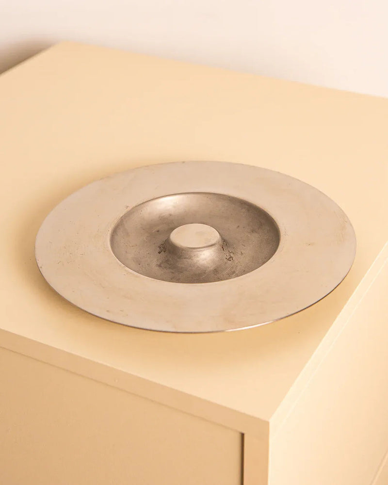 The Italian Circular Ashtray by Velca 80's from Treaptyque rests on a light-colored, rectangular surface, its round metallic design featuring a raised center that casts gentle shadows. This minimalistic ashtray embodies Italian craftsmanship with its modern and subtle aesthetic, infused with vintage 80's charm.