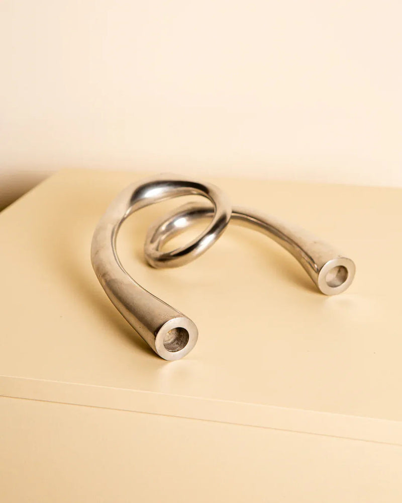 The Treaptyque Italian Candleholder in Cast Aluminum 80's, with its twisting design, sits elegantly on a beige surface against a cream backdrop. Its sleek, reflective finish and smooth curves embody minimalist elegance and modern aesthetic.
