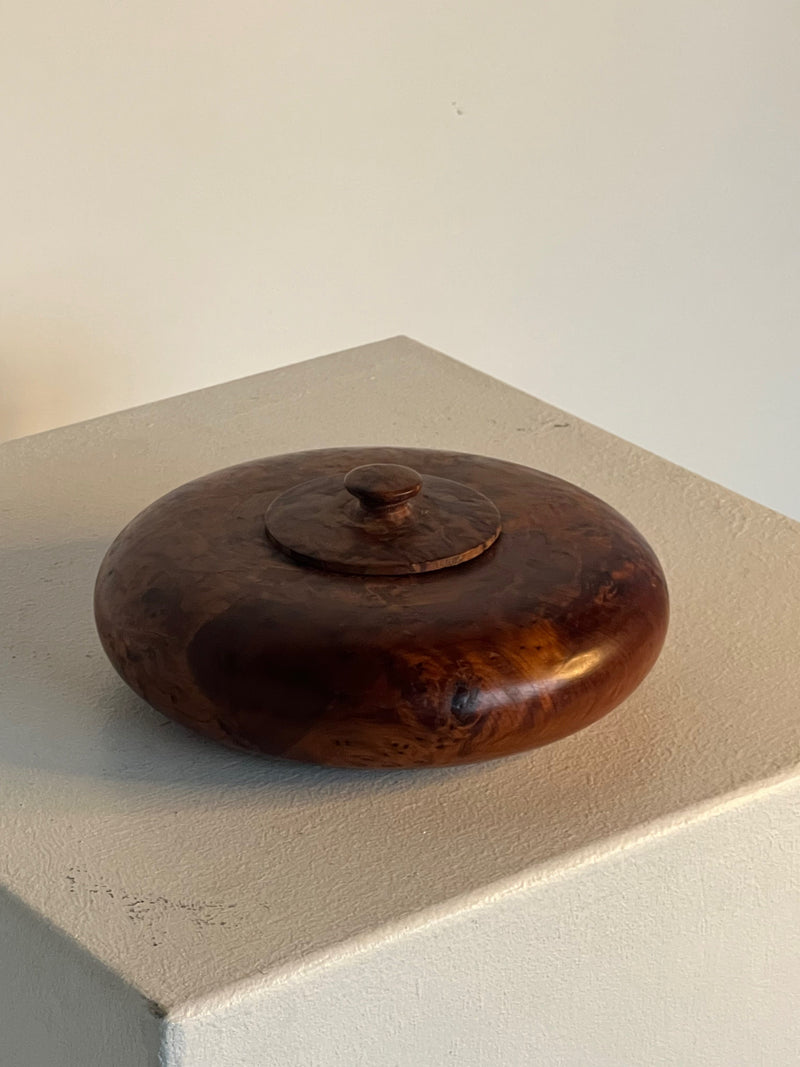 The Walnut Burl Shallow Bowl With Lid by Porter Studio is a beautifully crafted round dish featuring a small knob on top. Made from rich brown walnut burl, it showcases a smooth, glossy finish with visible grain patterns. Elegantly displayed on a white pedestal, the shallow bowl sits against a simple beige background.