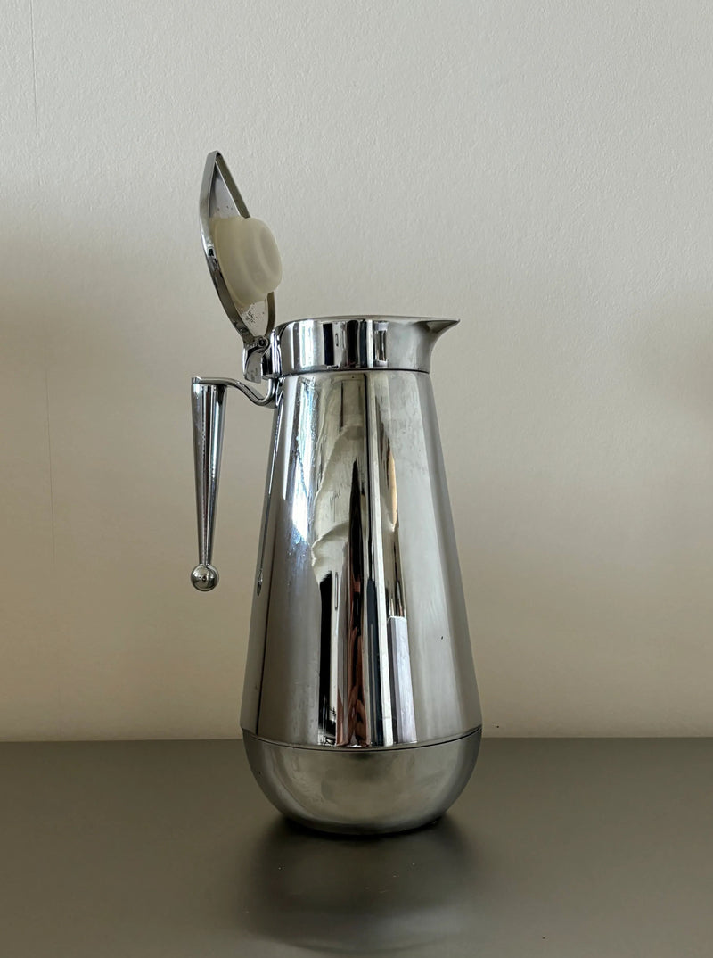 A polished silver CU-RA-TED Steel Kettle “Erhard Germany” in impeccable condition with a round base and an elongated spout sits on a gray surface against a plain white background. The kettle, reminiscent of an Erhard Germany design, features an open hinged lid and a cylindrical handle extending from its side.