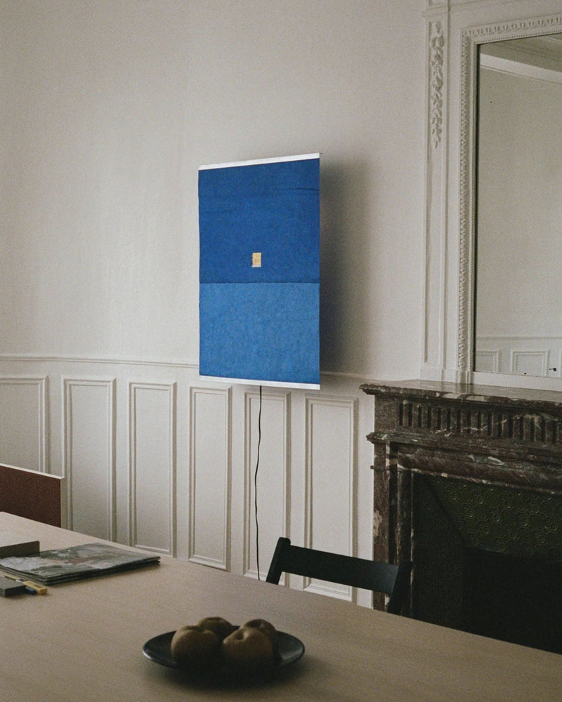 A minimalistic room with white paneled walls features a blue rectangular artwork above a dark fireplace. In the foreground, a wooden table holds a bowl of pears and a book, softly illuminated by natural light and the glow of TEGET's Panel Wall Lamp – Montenegro, handmade by Ana Kraš.