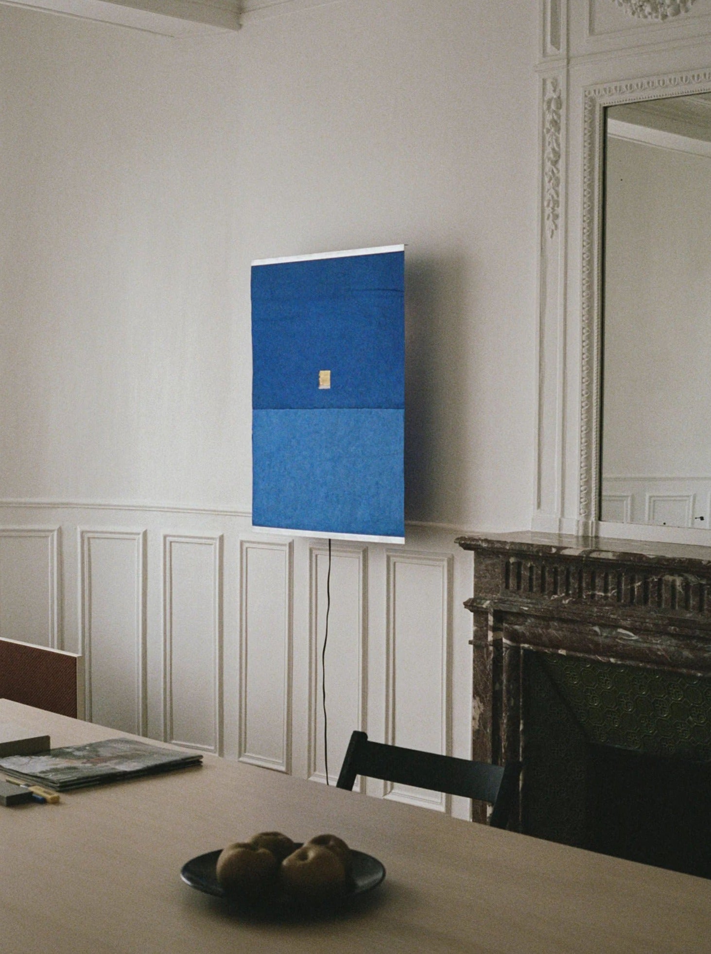 A minimalistic room with white paneled walls features a blue rectangular artwork above a dark fireplace. In the foreground, a wooden table holds a bowl of pears and a book, softly illuminated by natural light and the glow of TEGET's Panel Wall Lamp – Montenegro, handmade by Ana Kraš.