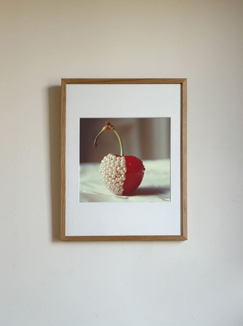 A framed fine art print on a white wall showcases a cherry dipped halfway into tiny white beads, resembling pearls. The cherry's stem curves gently over the fruit, creating a striking juxtaposition between the smooth red surface and the textured white half of this limited edition piece, "Cherry and Pearls" by Adeline Mai.