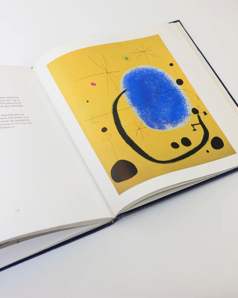 An open "Mirò The Masterworks Book" from Boga Avante Shop displays surreal abstract art with a large blue oval, black lines, small black shapes on a yellow background, and colored dots; the opposite page contains text.