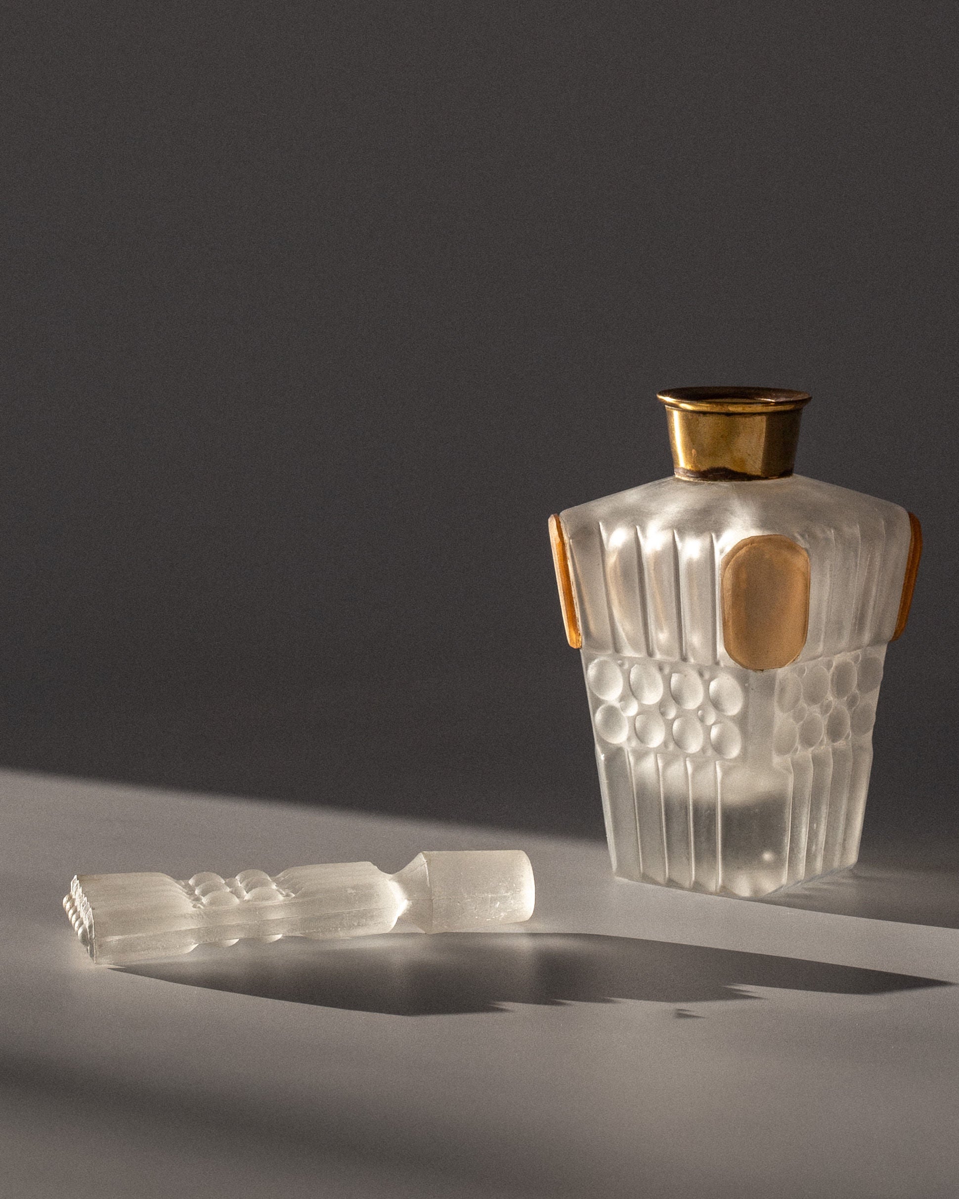 A Spigel Art Deco Perfume Bottle from France, 1930s, with geometric patterns and gold accents, rests on a surface. The frosted glass cap mirrors the design and lies beside it. Soft light highlights intricate details, casting elegant shadows.