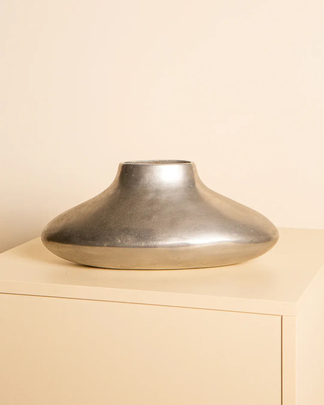 The 80's Cast Aluminium Vase by Treaptyque features a wide, flat, and rounded body with a narrow opening. It sits elegantly on a light tabletop against a beige backdrop, exuding modern Italian design influences.