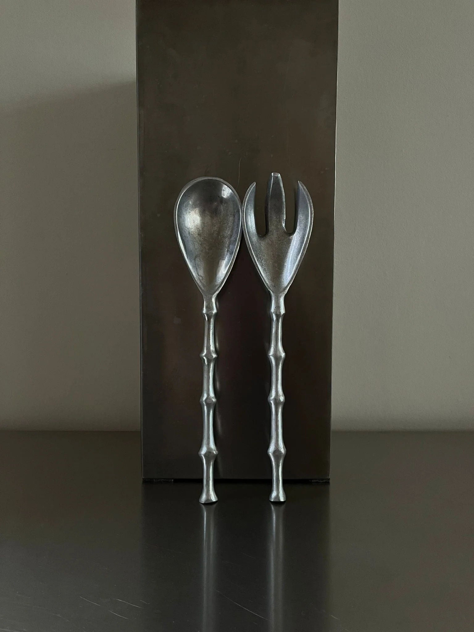 Two metallic serving utensils with handles designed to resemble bamboo. One utensil is a rounded spoon, and the other is a two-pronged fork. These silver-plated serving tools stand upright against a rectangular metallic block on a smooth surface. The background is neutral. This set of Lettuce Cutlery by CU-RA-TED adds an elegant touch to any dining experience.