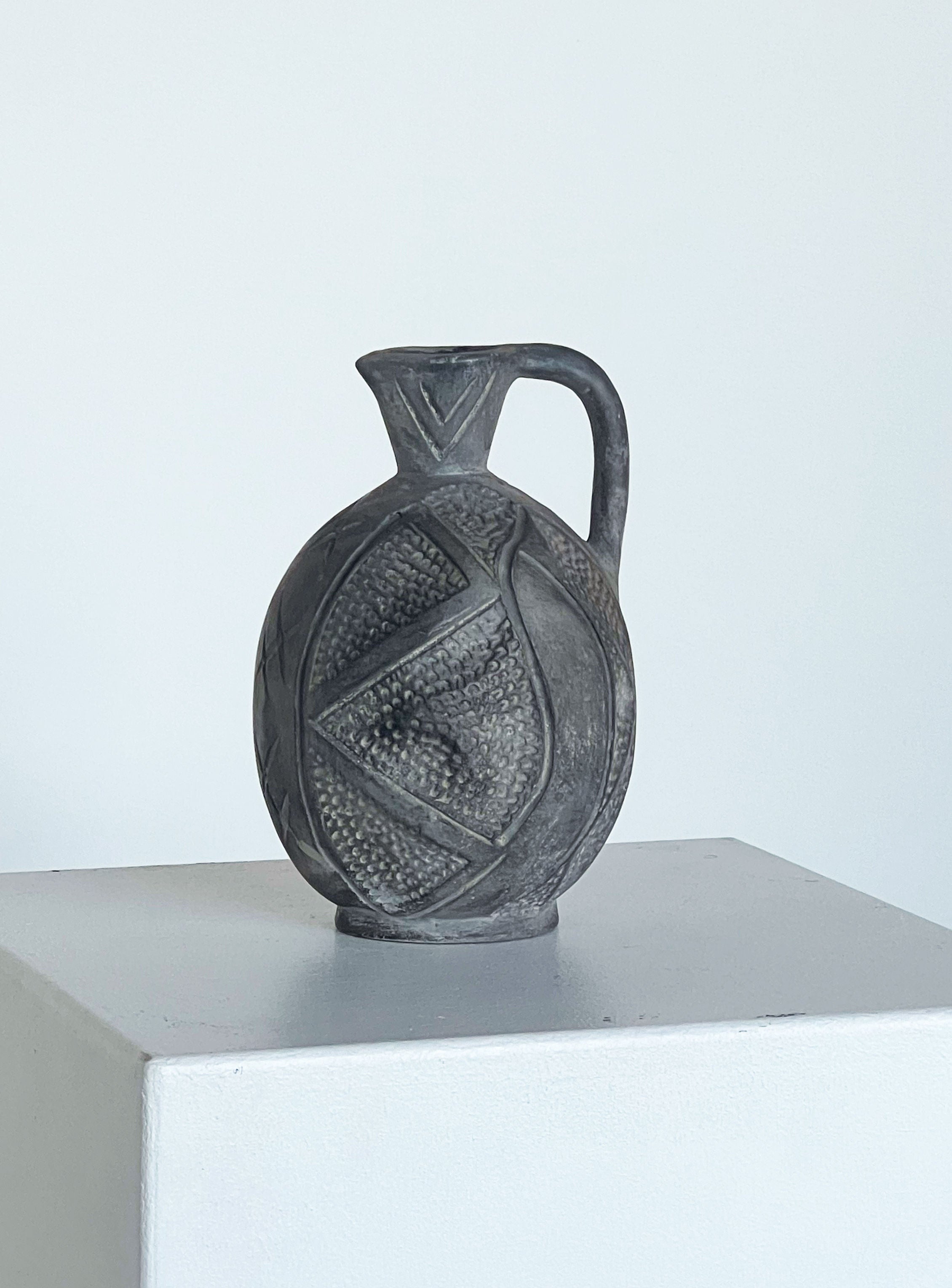 A handmade "Jug with Hand Markings" from Porter Studio, featuring an elegant round shape and a handle, sits on a white pedestal. It displays intricate embossed patterns and a narrow neck, with black and grey tones standing out against the plain, light gray background. This piece exemplifies artisan pottery at its finest.