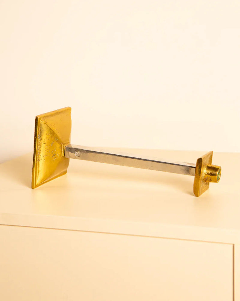 The Brutalist Candleholder by David Marshall, an 80's design from Treaptyque, presents a minimalist gold square base and lampshade, with a metallic stem that connects them for a sleek, modern aesthetic reminiscent of an elegant brass and silver candlestick on a light surface.