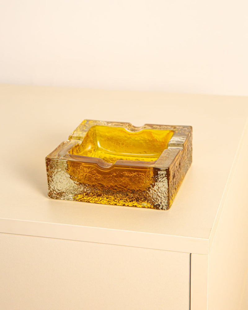The Square Ashtray in Italian glass 60s by Treaptyque, featuring textured amber glass and grooved edges, rests on a minimalist light surface against a plain backdrop, capturing classic 60s style.