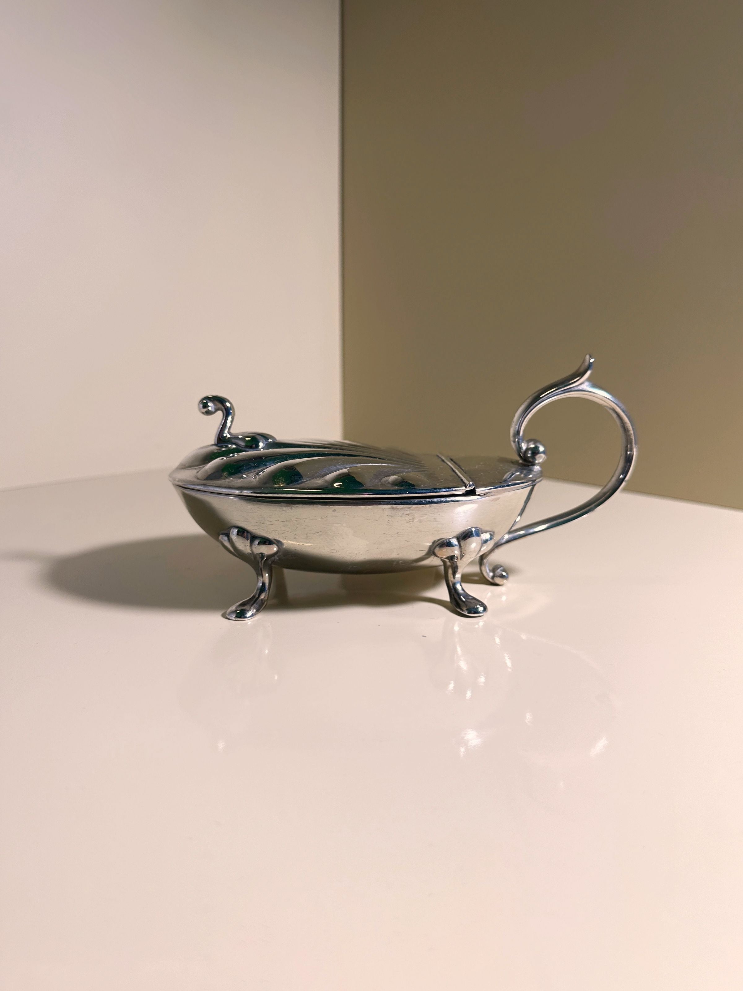 A Silver-plated Caviar Frog from the 1950s by Dodo Vintage, featuring a sleek curved handle and three ornate legs, sits on a glossy cream surface against a beige two-toned wall.