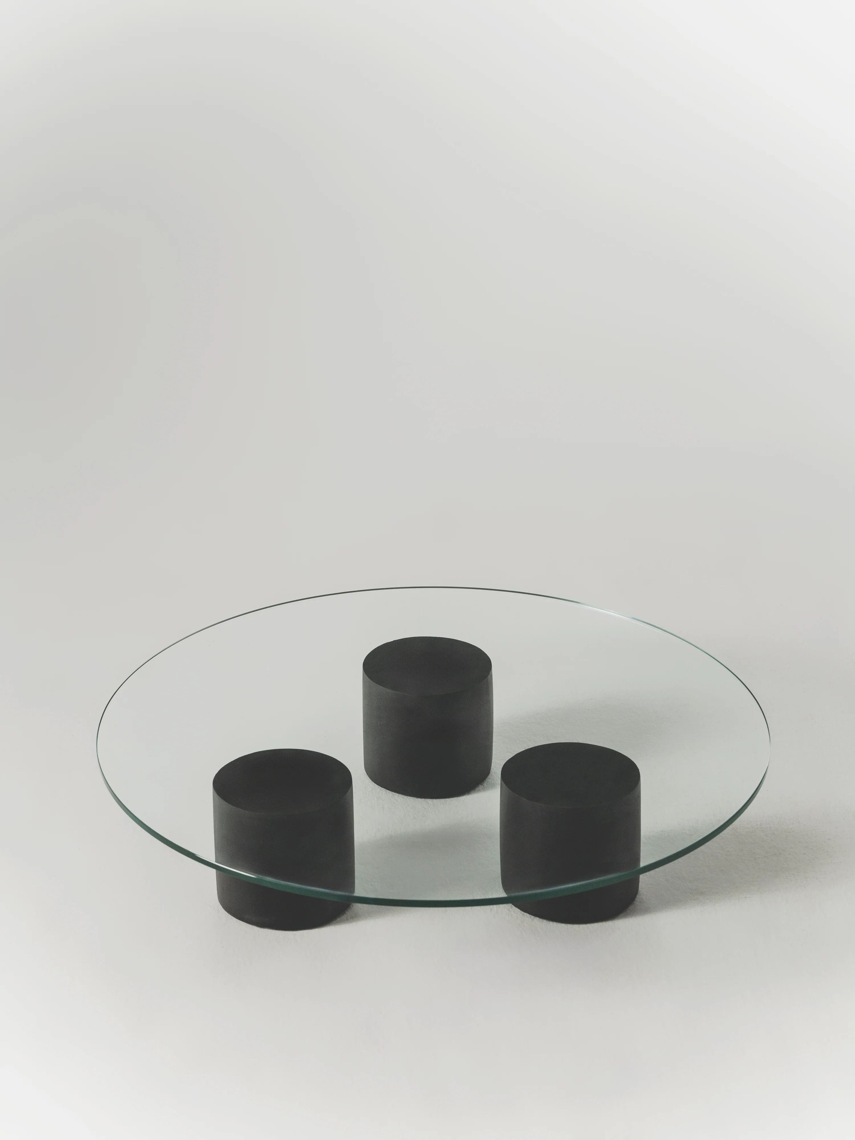 The Carmene Glass Coffee Table by MarlotBaus features a minimalist design with a round tempered glass top on three cylindrical black supports, set against a plain white background.