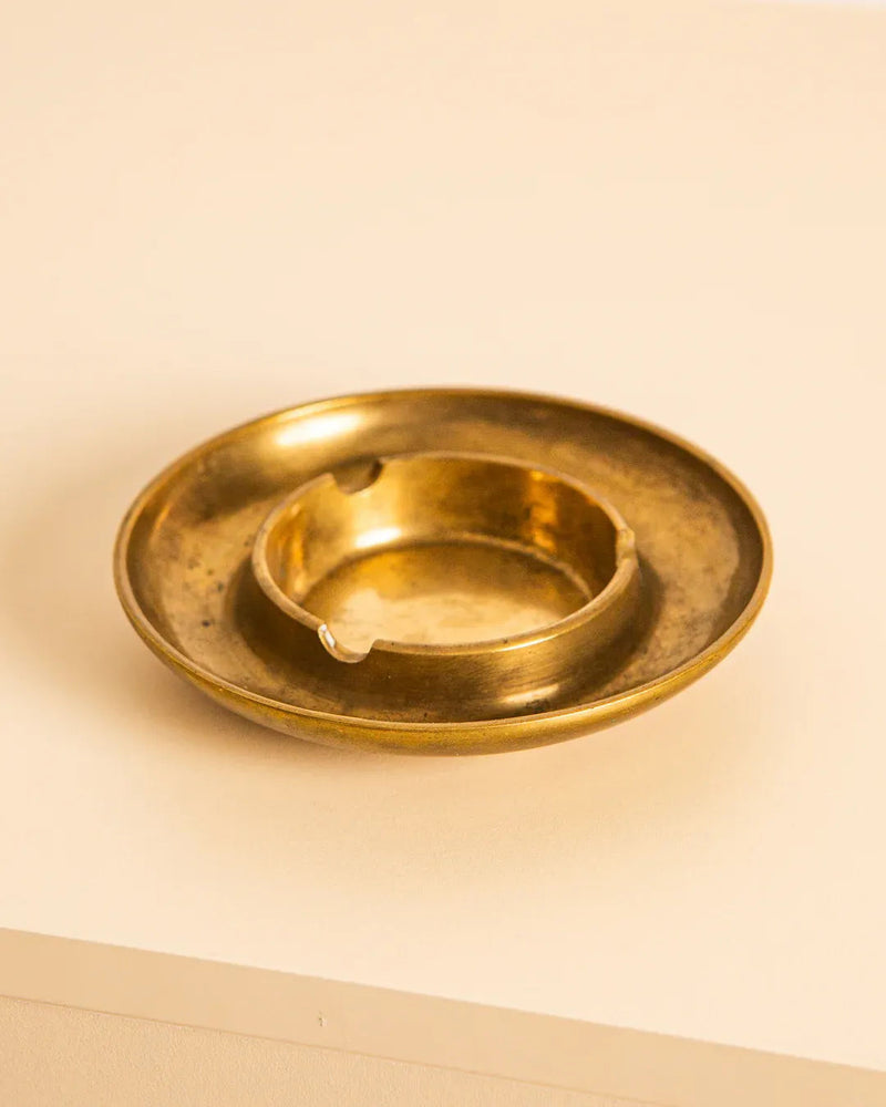 The Italian Circular Brass Ashtray 70's from Treaptyque, sitting on a light beige surface, exemplifies exquisite Italian craftsmanship. Its small circular design from the 70s includes a slightly raised center for holding cigarettes and displays a smooth, aged patina.