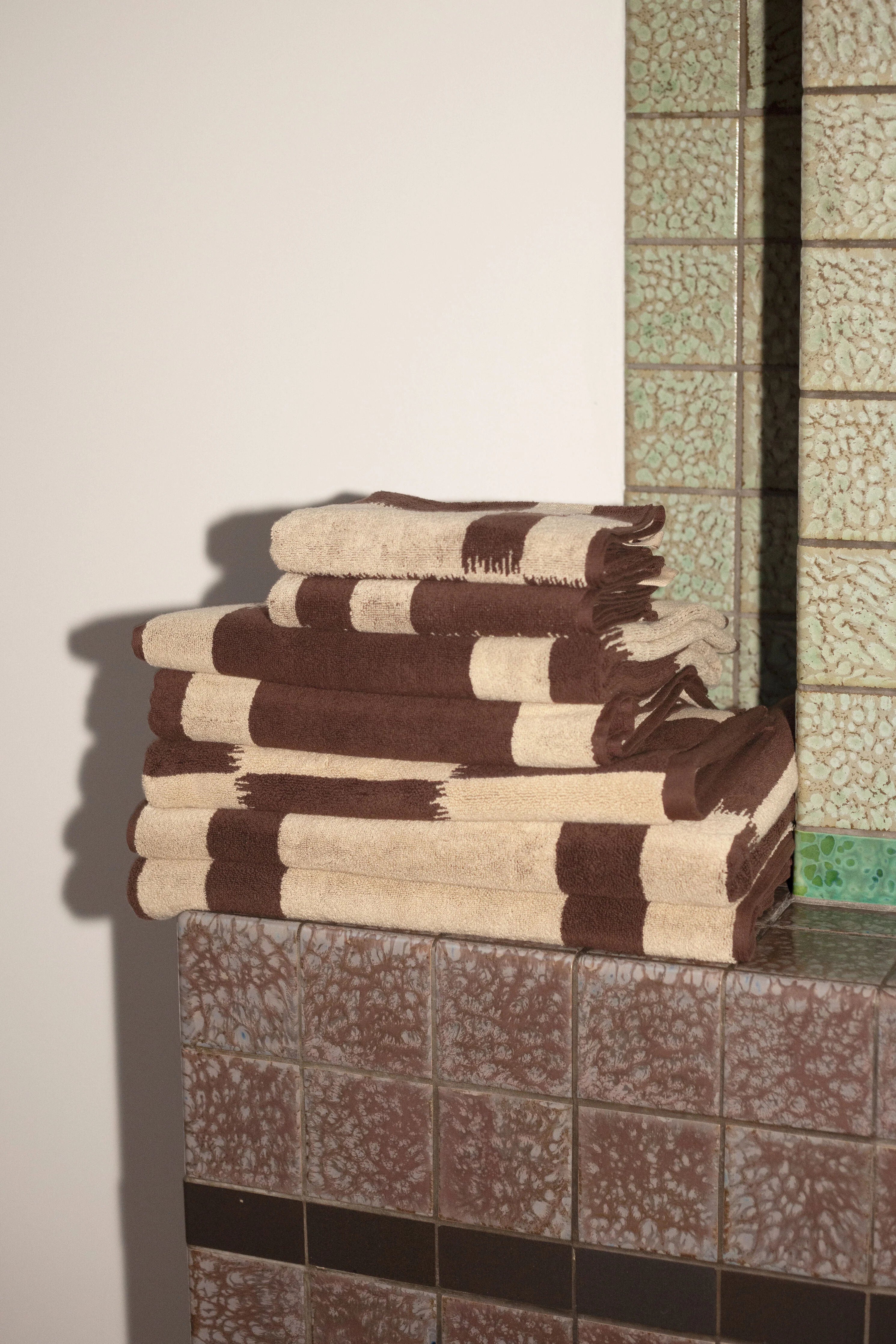 A stack of brown and beige striped towels placed on the ledge of a tiled wall with a green Ikat weaving pattern. Light casts a shadow on the textured surface, next to the Autumn Sonata Karin Bath Mat.