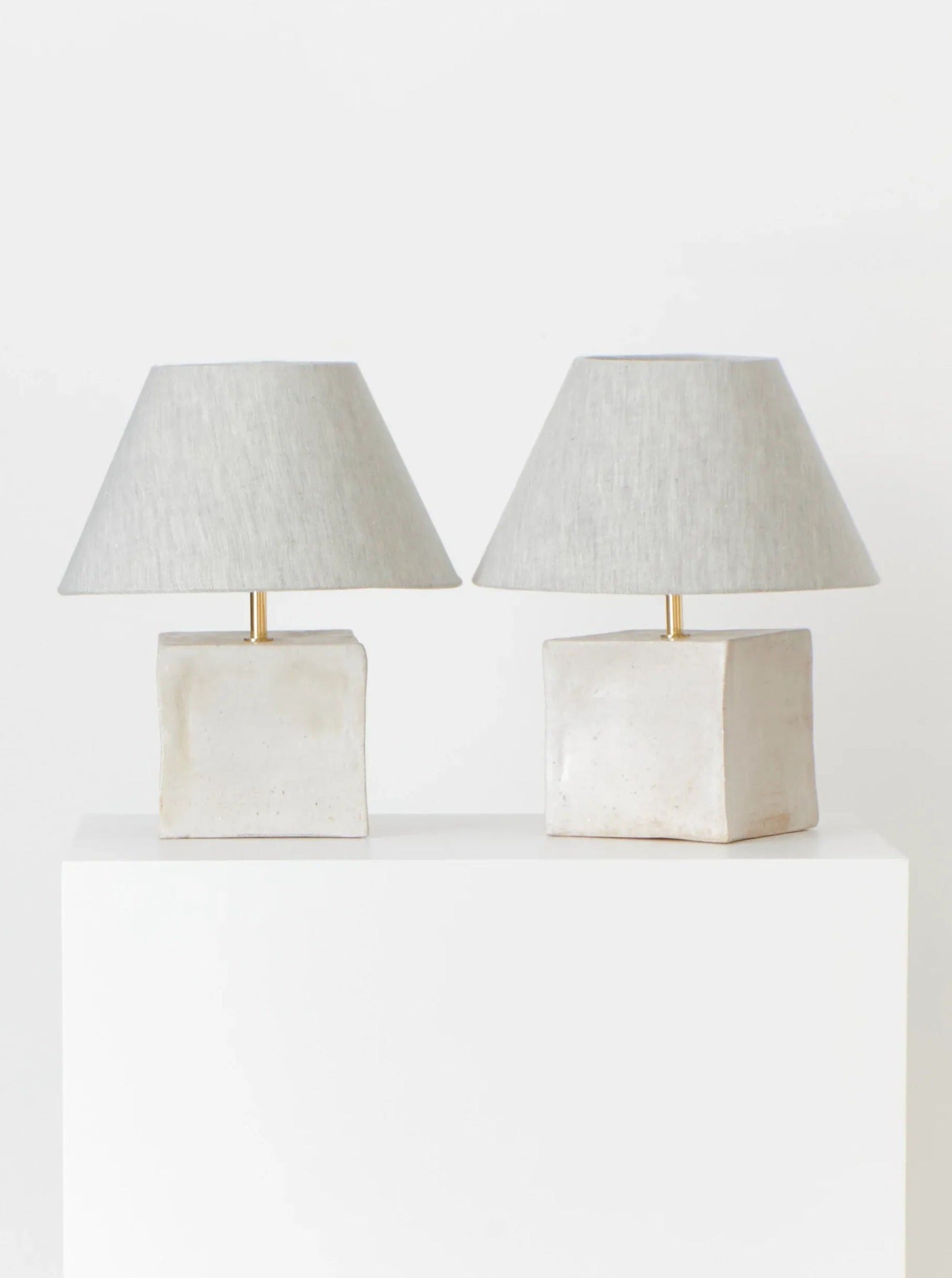 Two Arouca Table Lights by Project 213A with square, stone-textured bases and light gray fabric shades are displayed on a white rectangular pedestal against a plain white background. These artisanal table lights feature a minimalist design, emphasizing their clean lines and neutral tones.