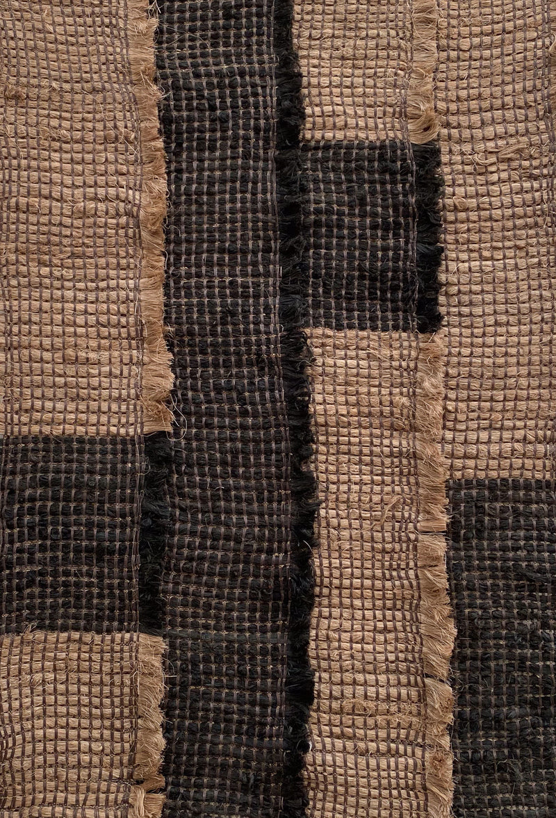 The image displays the Ink / Sand fabric by Sundance Studio, made from biodegradable banana fiber with a grid pattern. It shows dark and light brown stripes that create a rustic, woven look, with slightly frayed edges enhancing its natural charm.