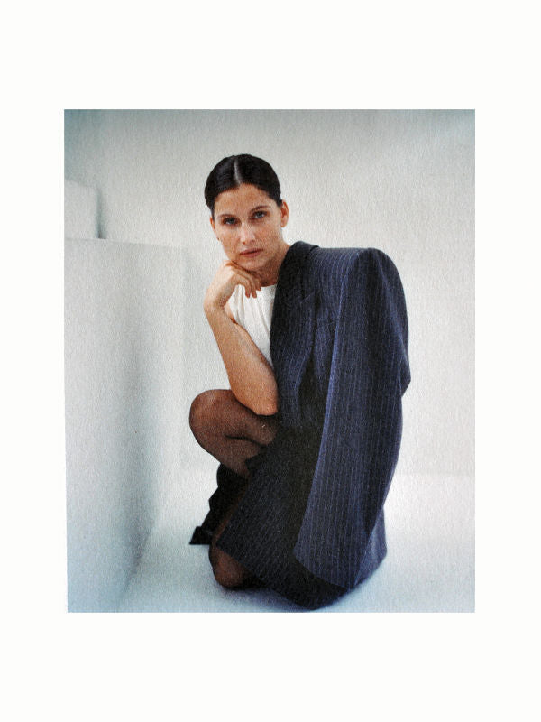 A woman with dark hair, tied back, gazes intently at the camera. She sits with one knee up, resting her elbow on her raised knee, and her chin on her hand. She wears a white top and drapes a blue pinstripe blazer over her shoulder. She exudes the confident elegance of a Marfa Muse by Maison Plage against a minimalist background.