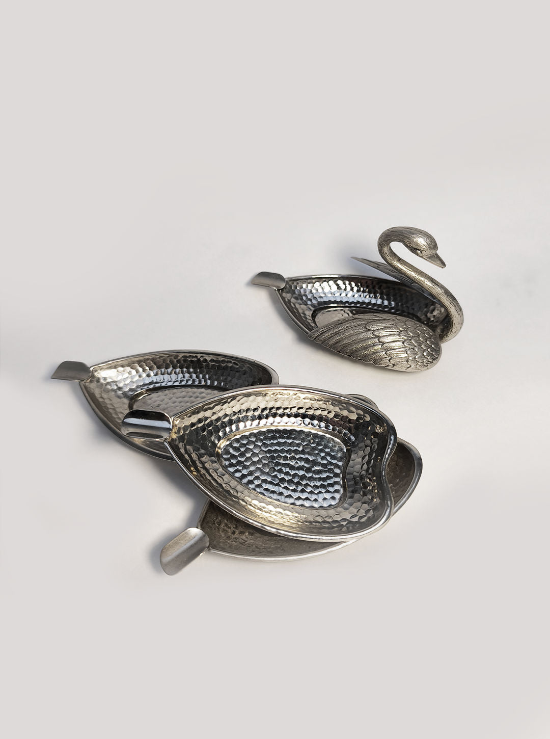 The Swan Ashtray set by Les Objoies features three intricately designed silver bowls, resembling elegant swans, that form a chic metallic centerpiece. Two of the bowls nest together gracefully, while the third stands independently. Each bowl is adorned with a hammered texture and has a graceful curved neck, offering an elegant alternative for smoking areas.