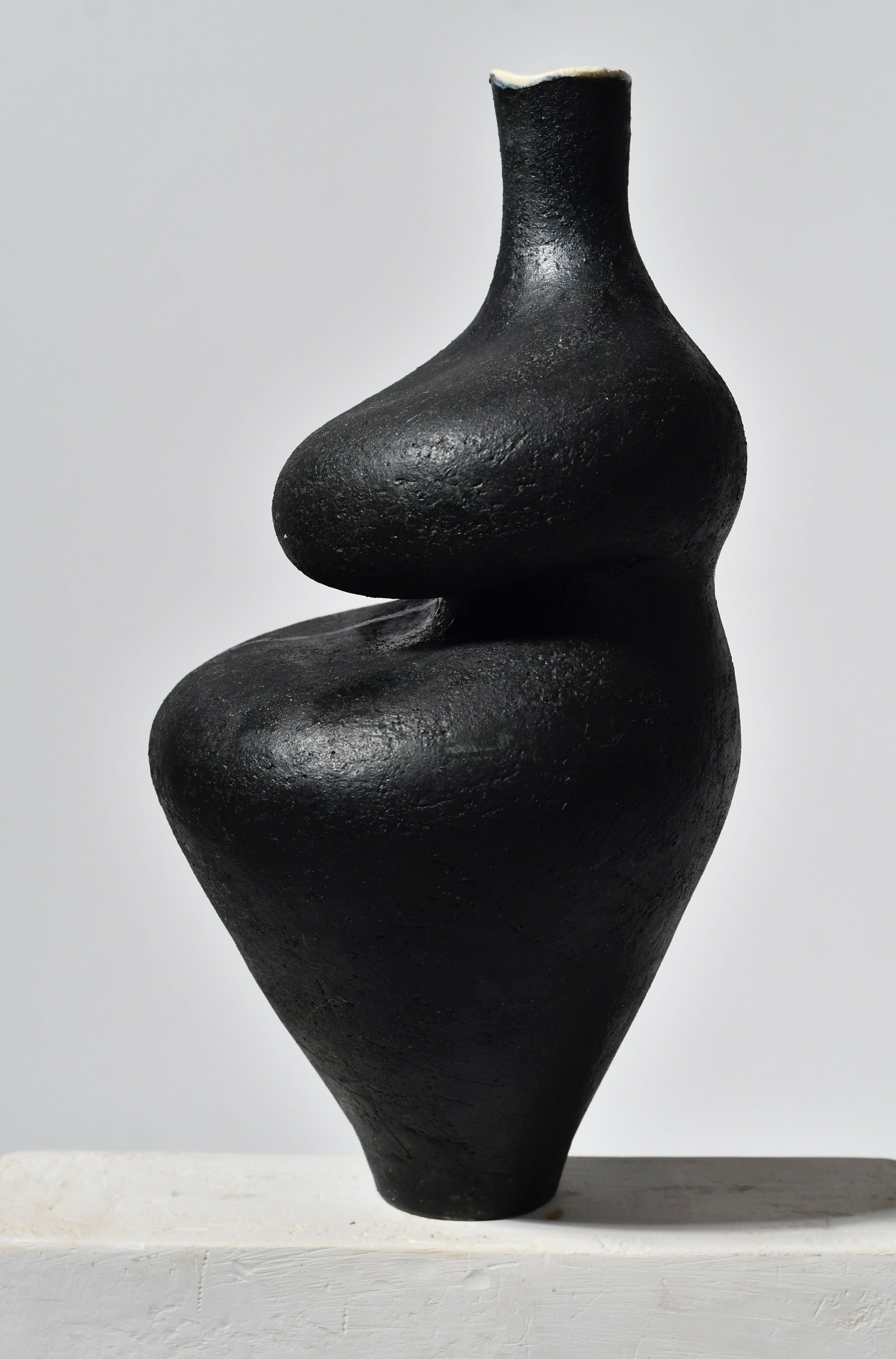 A sculptural Black Lilith with Oxide Drip ceramic vase by Lucia Mondadori, with a unique abstract form resembling intertwined human figures, displayed against a white background, features a subtle oxide drip.