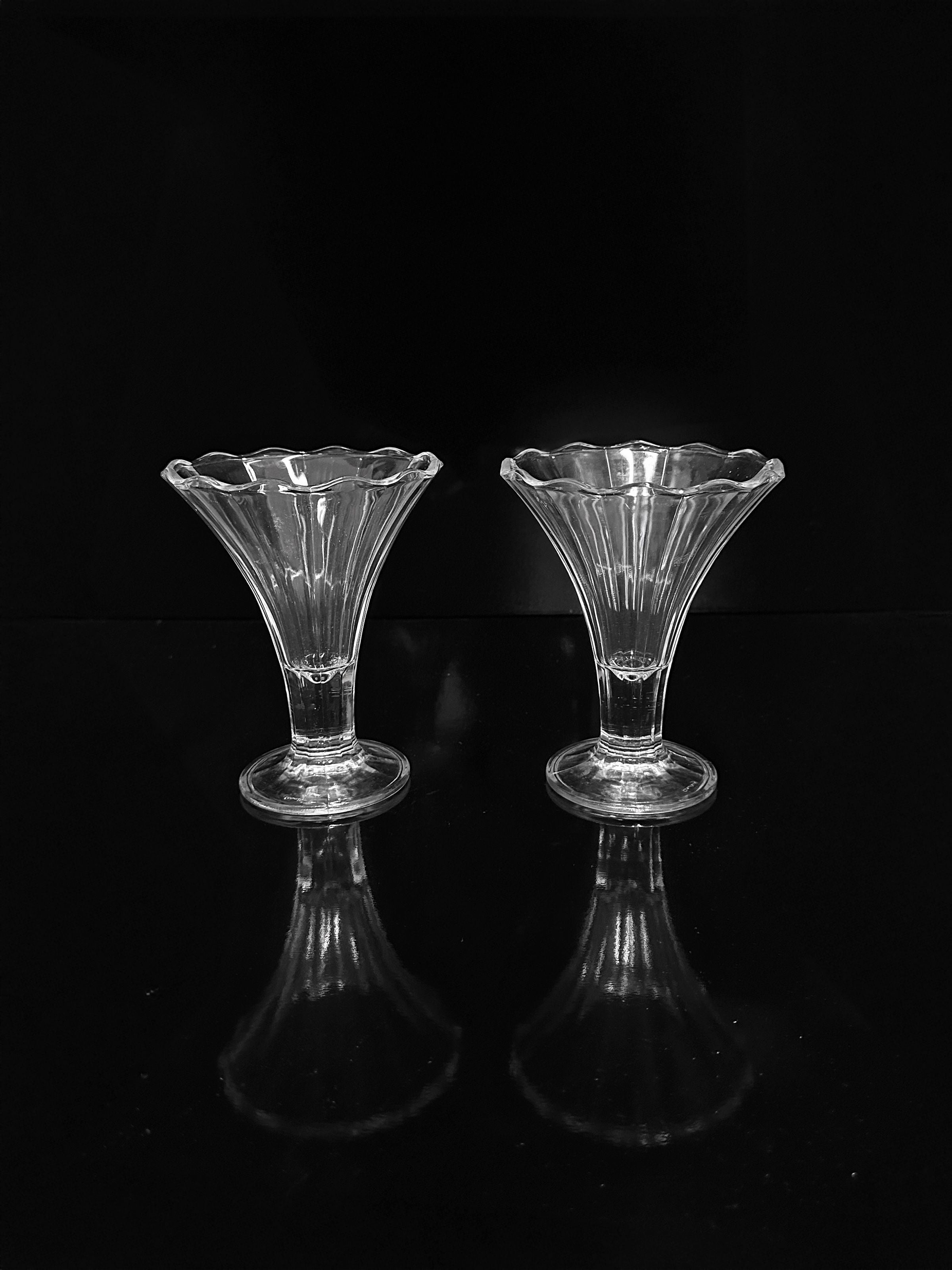 Two stylish Italian Art Deco Cocktail Glasses from Dodo Vintage, featuring clear glass with flared, scalloped rims reminiscent of elegant ice cream cups, are placed side by side against a black background. Their reflections shimmer on the glossy surface beneath them.